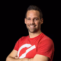 Lächelnder Mann mit kurzen braunen Haaren, Bart und rotem T-Shirt mit weißem Logo, vor schwarzem Hintergrund.