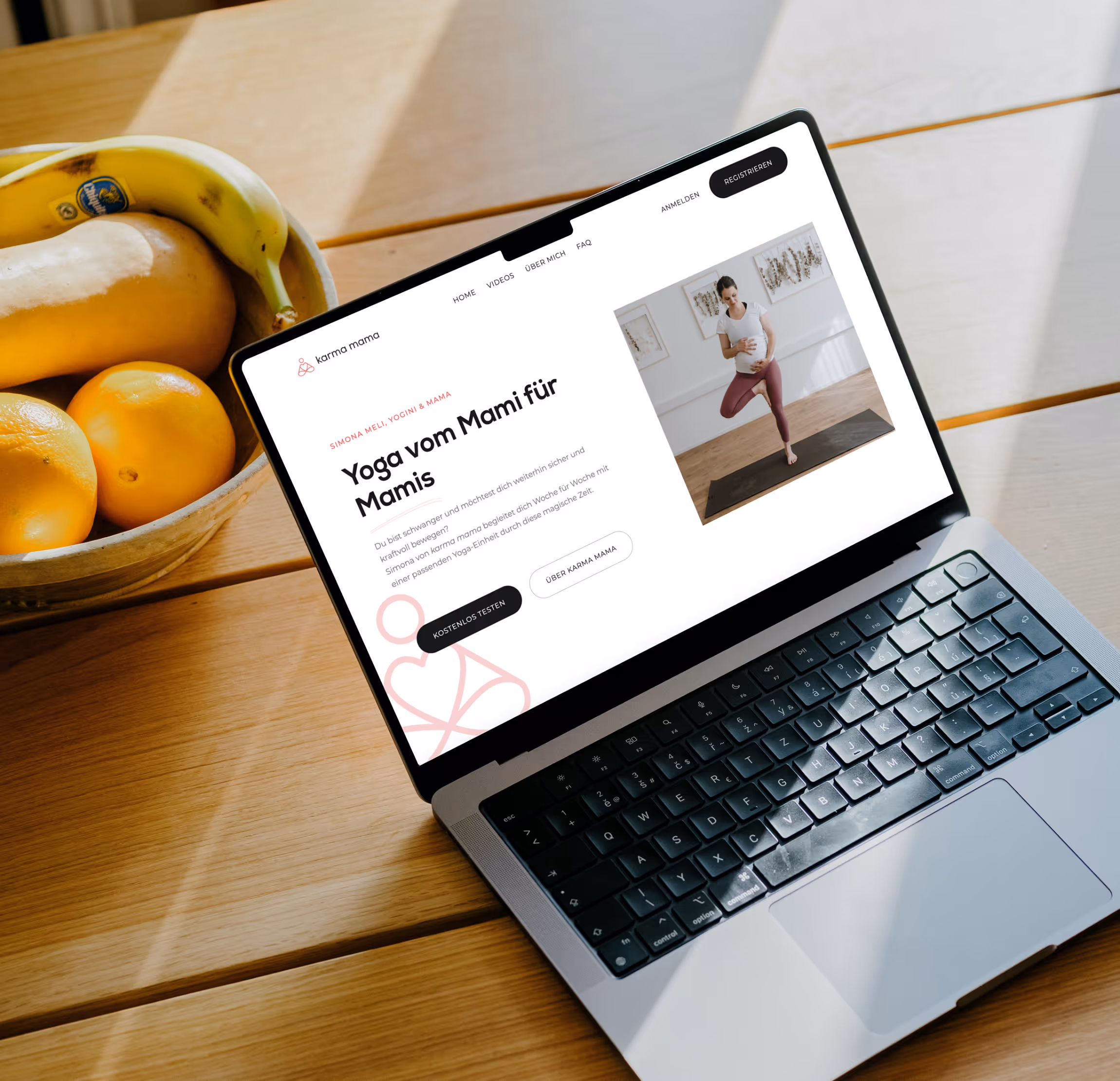 Ein MacBook auf einem Holztisch zeigt eine Webseite für Yoga von Müttern, daneben eine Schale mit Bananen und Orangen.