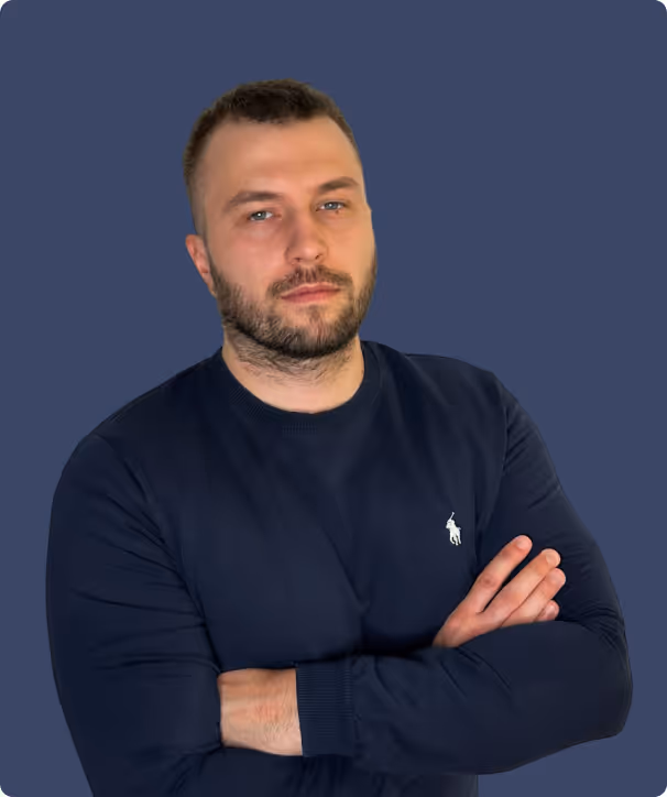 Selbstbewusster Mann mit kurzem Haar und Bart, verschränkten Armen, trägt ein dunkelblaues Langarmshirt vor blauem Hintergrund.
