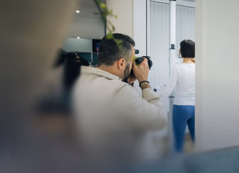 Ein Mann fotografiert mit Kamera eine Frau, die vor einem Fenster mit weißen Jalousien steht.