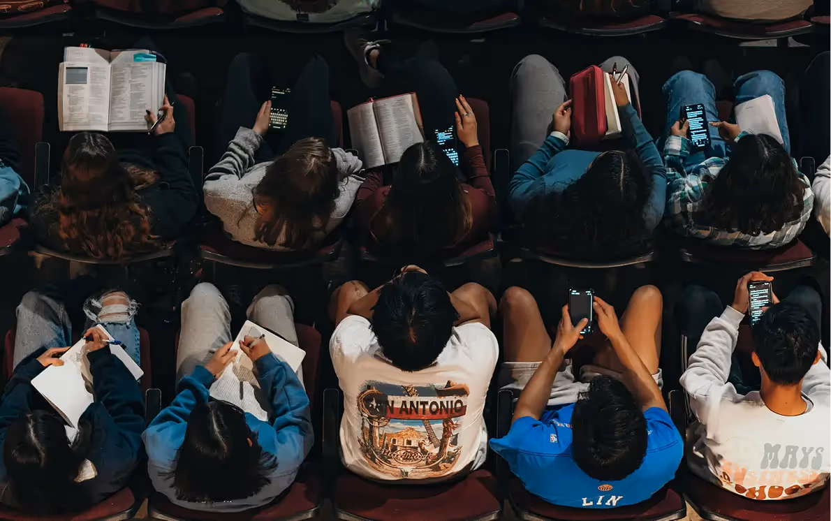 Students sitting in a lecture hall, some taking notes while others use their phones.