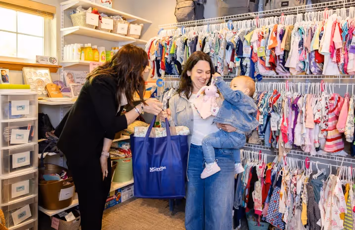 Two women smiling while exchanging a Care Net tote bag in a room filled with baby clothes and supplies, with one holding a baby.