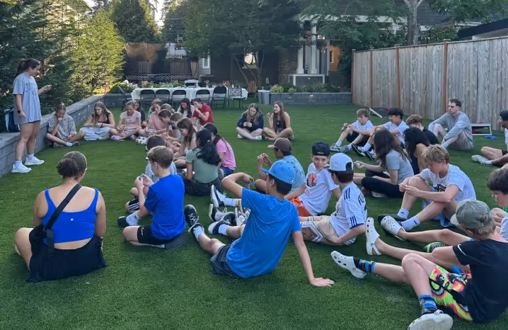 Group of teenagers sitting on a lawn during a youth gathering, listening to a speaker at the front on a summer afternoon.