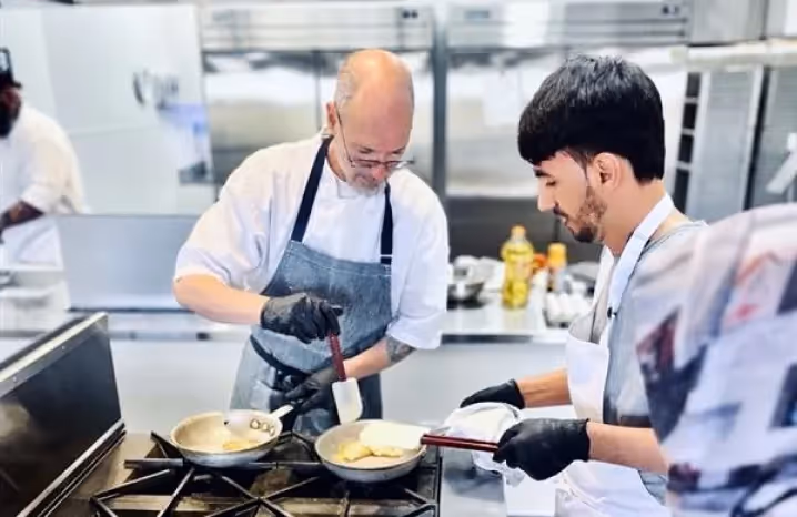 Chef instructing a student on how to flip food in a pan during a cooking class in a professional kitchen.