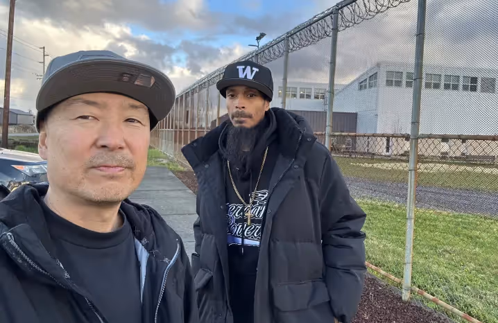 Two men standing outside a fenced facility, dressed warmly in jackets and caps, appearing to visit or work near a correctional institution.