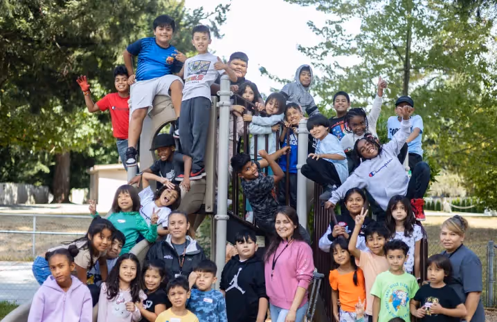 Group of children and adults smiling and posing together on a playground structure surrounded by trees.