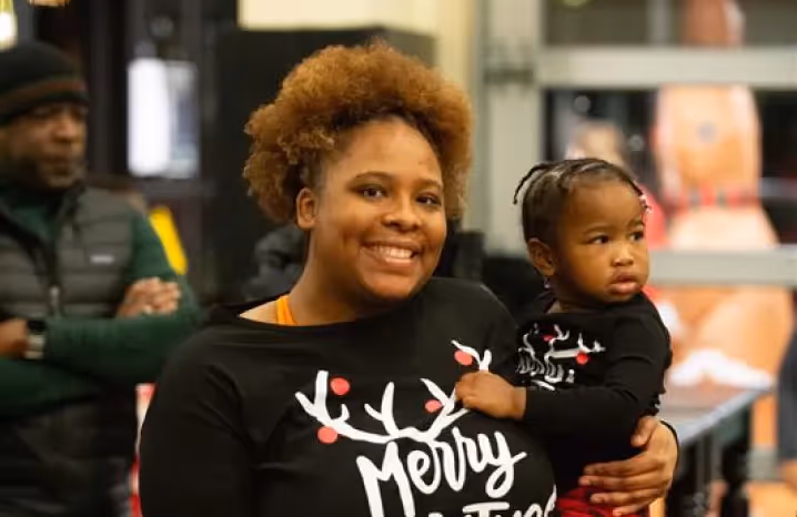 Smiling woman holding her young daughter while wearing matching festive Christmas shirts at a holiday event.