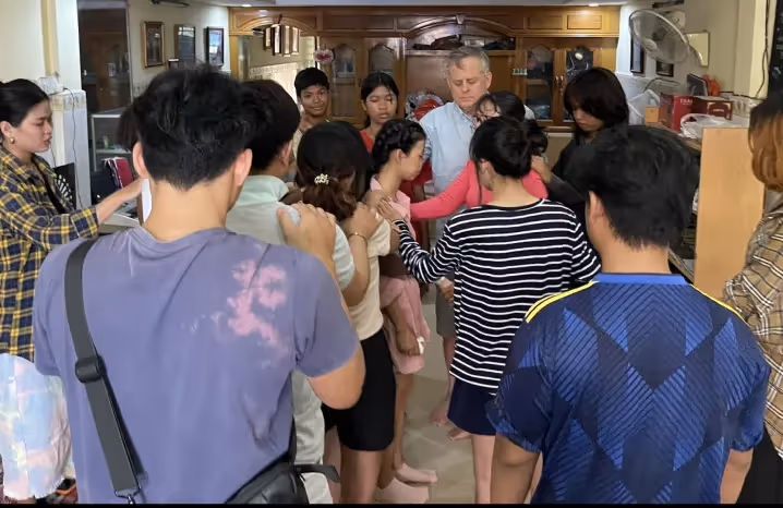 Group of people gathered in a room, standing close together and praying with hands on one another.