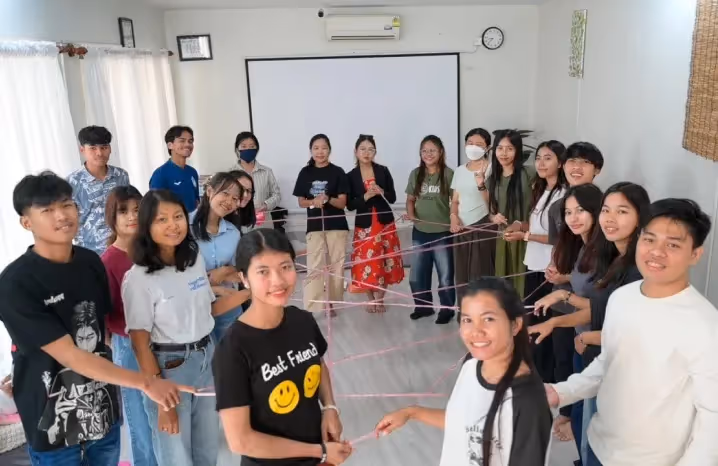 Young adults participating in a team-building activity using strings, forming a web while smiling in a classroom.