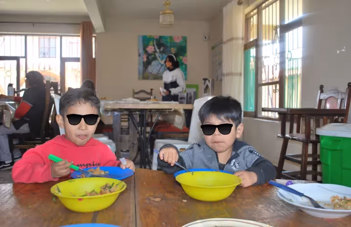 Two young children wearing sunglasses sitting at a table, eating meals from yellow bowls inside a brightly lit room.