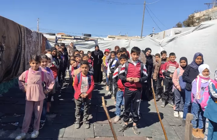 Children standing in lines outdoors between tents in a refugee camp, smiling and looking toward the camera.