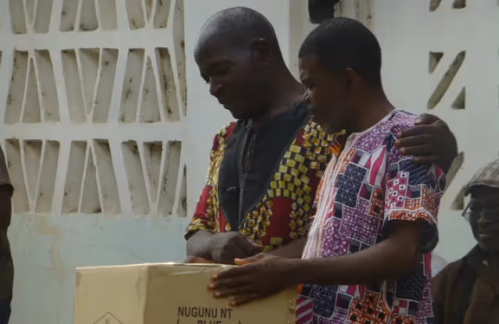 Two men standing outside a building, smiling and holding a box marked “Nugunu NT Bible,” suggesting a Bible distribution event.