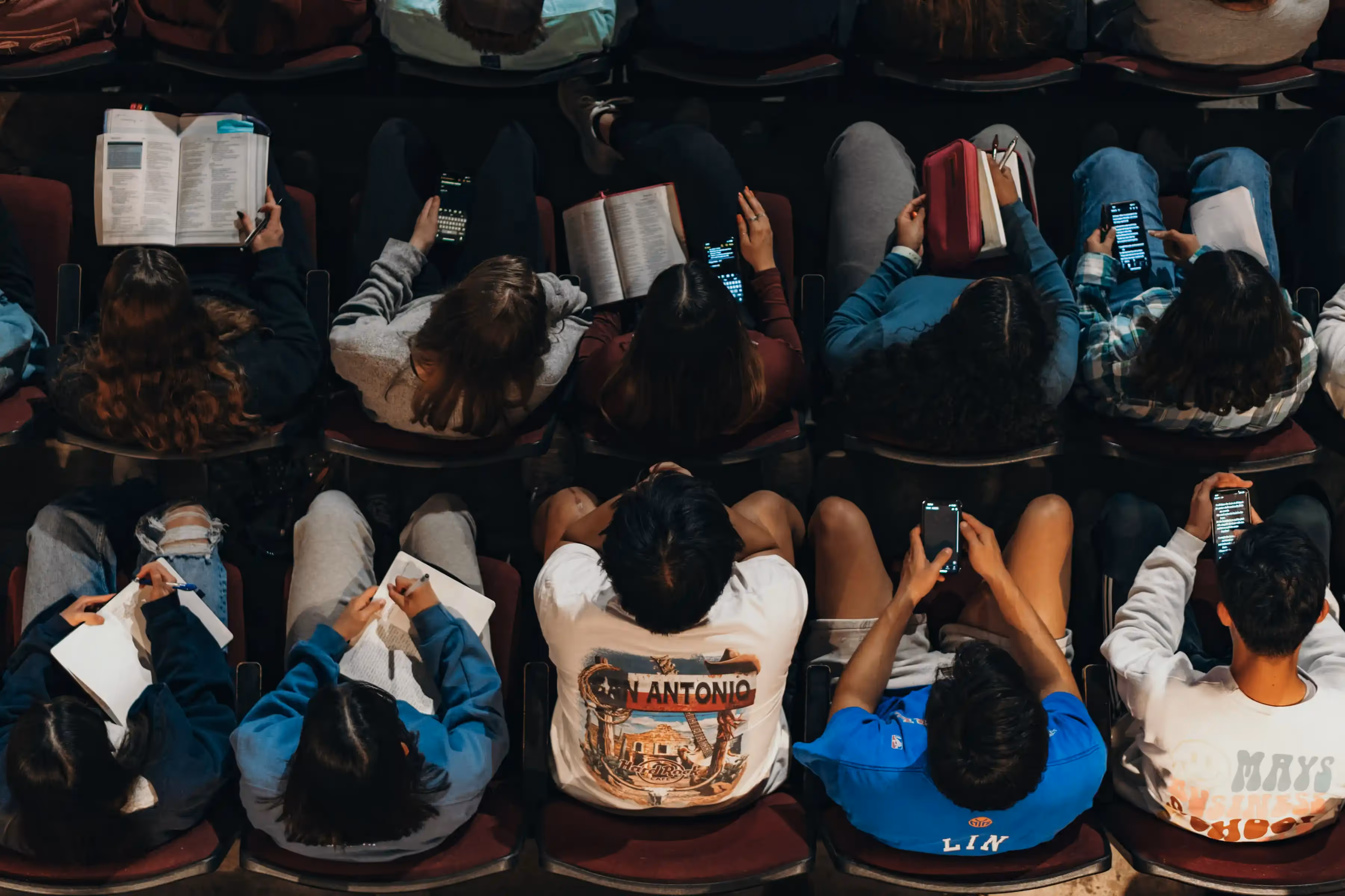 Group of students sitting in rows taking notes and reading from phones and Bibles during a study session. 