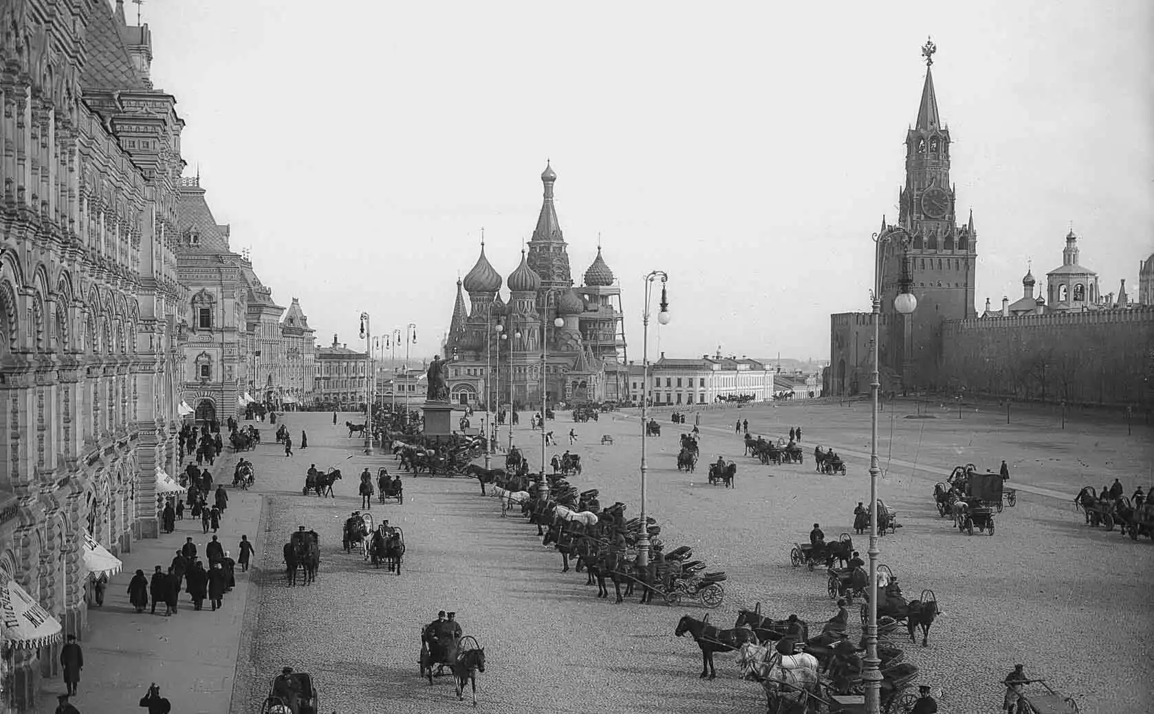 Vista aérea do conjunto arquitetônico da Praça Vermelha em Moscou sob um céu claro com nuvens. No centro, sobre uma elevação, fica a Catedral da Intercessão da Santíssima Mãe de Deus sobre o Fosso, conhecida como Catedral de São Basílio, com cúpulas retorcidas brilhantes em verde, vermelho e amarelo. À esquerda, estende-se em ângulo a longa muralha de tijolos vermelhos do Kremlin de Moscou com a alta Torre Spasskaya, que possui um relógio circular. À direita, estende-se a ampla esplanada cinza e vazia da Praça Vermelha, ao fim da qual, ao fundo, avista-se o edifício vermelho-escuro do Museu Histórico Estatal.