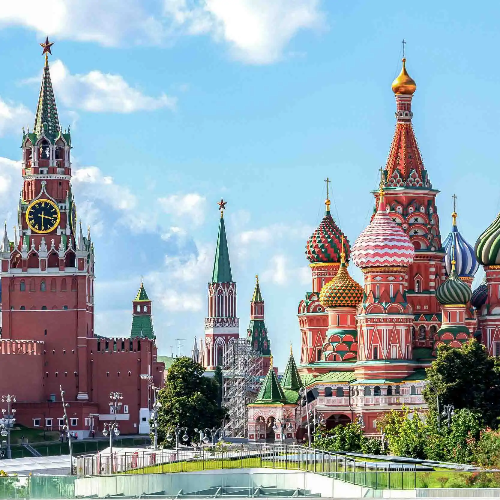 Conjunto histórico e arquitetônico da Praça Vermelha em Moscou. À direita ergue-se a Catedral da Intercessão da Santíssima Mãe de Deus sobre o Fosso, conhecida como Catedral de São Basílio, de tijolos vermelhos com nove igrejas em forma de torre e cúpulas bulbosas coloridas com padrões em verde, vermelho, amarelo e azul. À esquerda está a Torre Spasskaya do Kremlin de Moscou com detalhes em pedra branca, encimada por uma tenda verde com uma estrela de rubi brilhante do Kremlin. Na fachada da torre, vê-se o relógio carrilhão circular preto da Torre Spasskaya com números dourados. No centro aparecem partes da Torre Nabatnaya e da Torre Tsarskaya, com o céu azul ao fundo.
