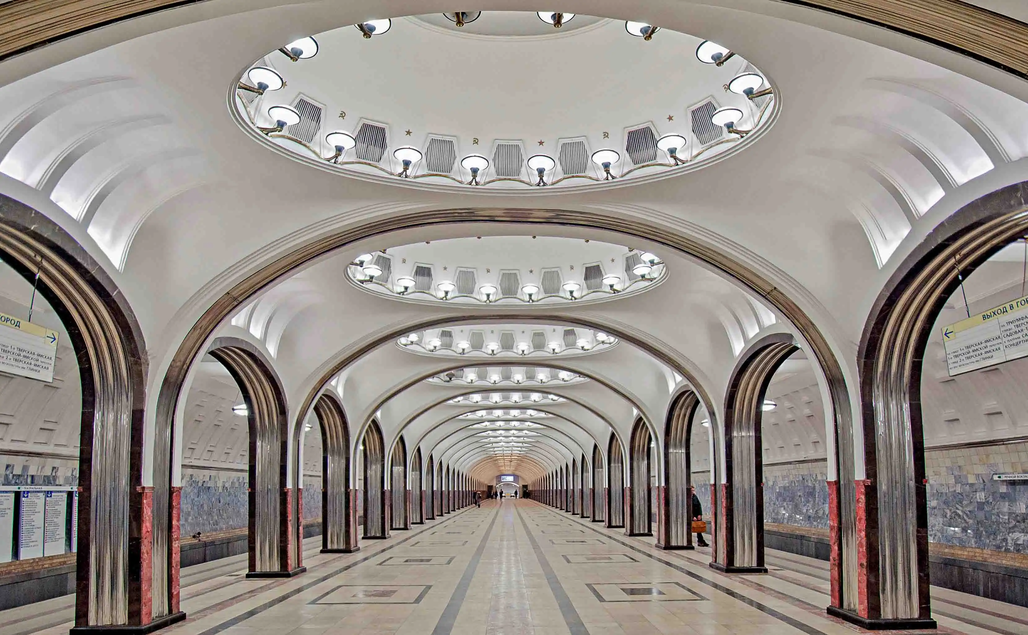 Conjunto histórico e arquitetônico da Praça Vermelha em Moscou. À direita ergue-se a Catedral da Intercessão da Santíssima Mãe de Deus sobre o Fosso, conhecida como Catedral de São Basílio, de tijolos vermelhos com nove igrejas em forma de torre e cúpulas bulbosas coloridas com padrões em verde, vermelho, amarelo e azul. À esquerda está a Torre Spasskaya do Kremlin de Moscou com detalhes em pedra branca, encimada por uma tenda verde com uma estrela de rubi brilhante do Kremlin. Na fachada da torre, vê-se o relógio carrilhão circular preto da Torre Spasskaya com números dourados. No centro aparecem partes da Torre Nabatnaya e da Torre Tsarskaya, com o céu azul ao fundo.