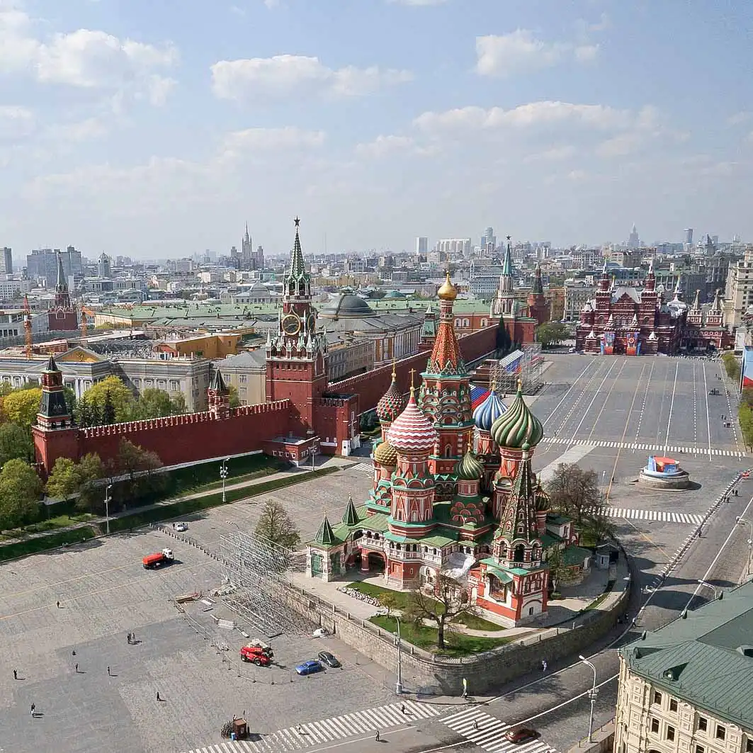 Vista aérea do conjunto arquitetônico da Praça Vermelha em Moscou sob um céu claro com nuvens. No centro, sobre uma elevação, fica a Catedral da Intercessão da Santíssima Mãe de Deus sobre o Fosso, conhecida como Catedral de São Basílio, com cúpulas retorcidas brilhantes em verde, vermelho e amarelo. À esquerda, estende-se em ângulo a longa muralha de tijolos vermelhos do Kremlin de Moscou com a alta Torre Spasskaya, que possui um relógio circular. À direita, estende-se a ampla esplanada cinza e vazia da Praça Vermelha, ao fim da qual, ao fundo, avista-se o edifício vermelho-escuro do Museu Histórico Estatal.