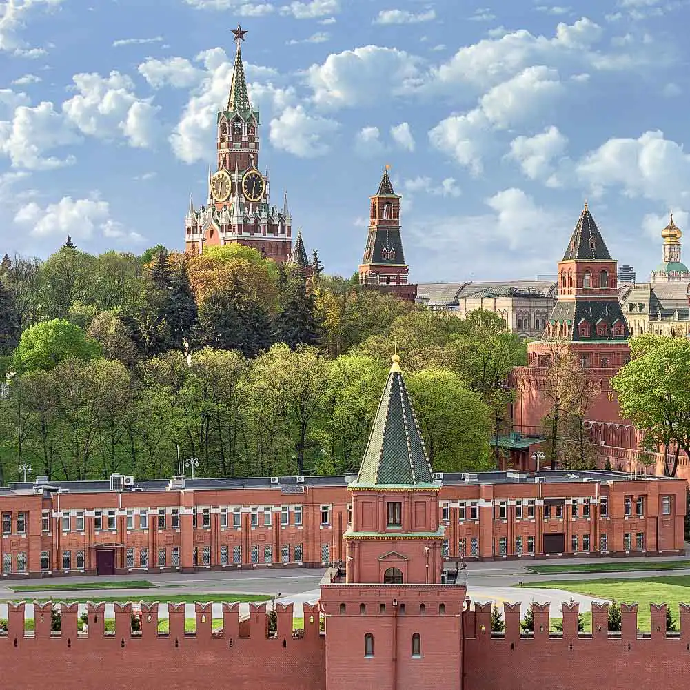 Fragmento do complexo fortificado do Kremlin de Moscou em um dia ensolarado. Em primeiro plano, acima da muralha de tijolos com ameias, ergue-se a robusta e quadrada Torre Taynitskaya com um teto de tenda verde. Atrás dela espalha-se um denso bosque verde, do qual sobressai ao fundo a alta Torre Spasskaya com o seu relógio carrilhão e uma estrela de rubi no pináculo, além da mais esguia Torre do Senado. À direita, avista-se parte da Torre Konstantino-Eleninskaya e a cúpula de uma catedral, com o céu coberto por nuvens cúmulas leves.