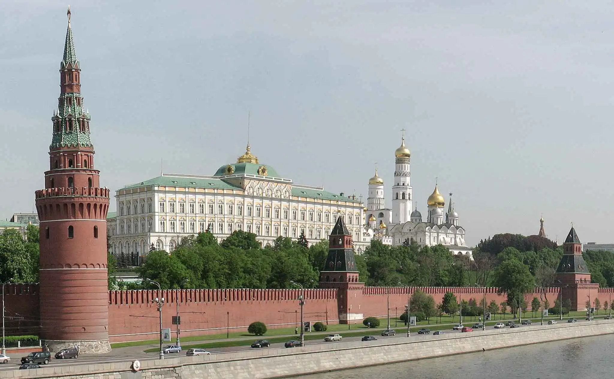 Fragmento do complexo fortificado do Kremlin de Moscou em um dia ensolarado. Em primeiro plano, acima da muralha de tijolos com ameias, ergue-se a robusta e quadrada Torre Taynitskaya com um teto de tenda verde. Atrás dela espalha-se um denso bosque verde, do qual sobressai ao fundo a alta Torre Spasskaya com o seu relógio carrilhão e uma estrela de rubi no pináculo, além da mais esguia Torre do Senado. À direita, avista-se parte da Torre Konstantino-Eleninskaya e a cúpula de uma catedral, com o céu coberto por nuvens cúmulas leves.
