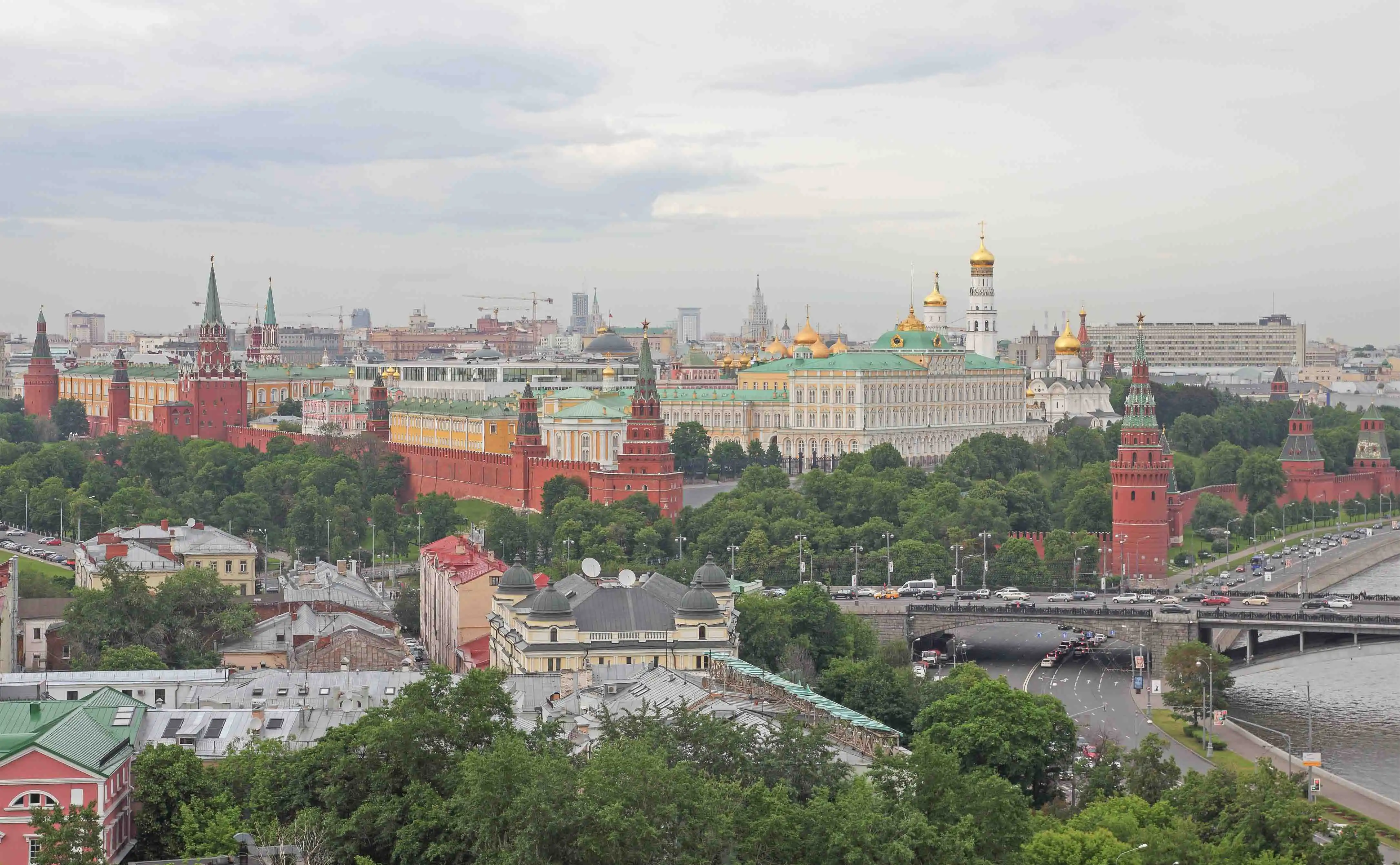Fragmento do complexo fortificado do Kremlin de Moscou em um dia ensolarado. Em primeiro plano, acima da muralha de tijolos com ameias, ergue-se a robusta e quadrada Torre Taynitskaya com um teto de tenda verde. Atrás dela espalha-se um denso bosque verde, do qual sobressai ao fundo a alta Torre Spasskaya com o seu relógio carrilhão e uma estrela de rubi no pináculo, além da mais esguia Torre do Senado. À direita, avista-se parte da Torre Konstantino-Eleninskaya e a cúpula de uma catedral, com o céu coberto por nuvens cúmulas leves.