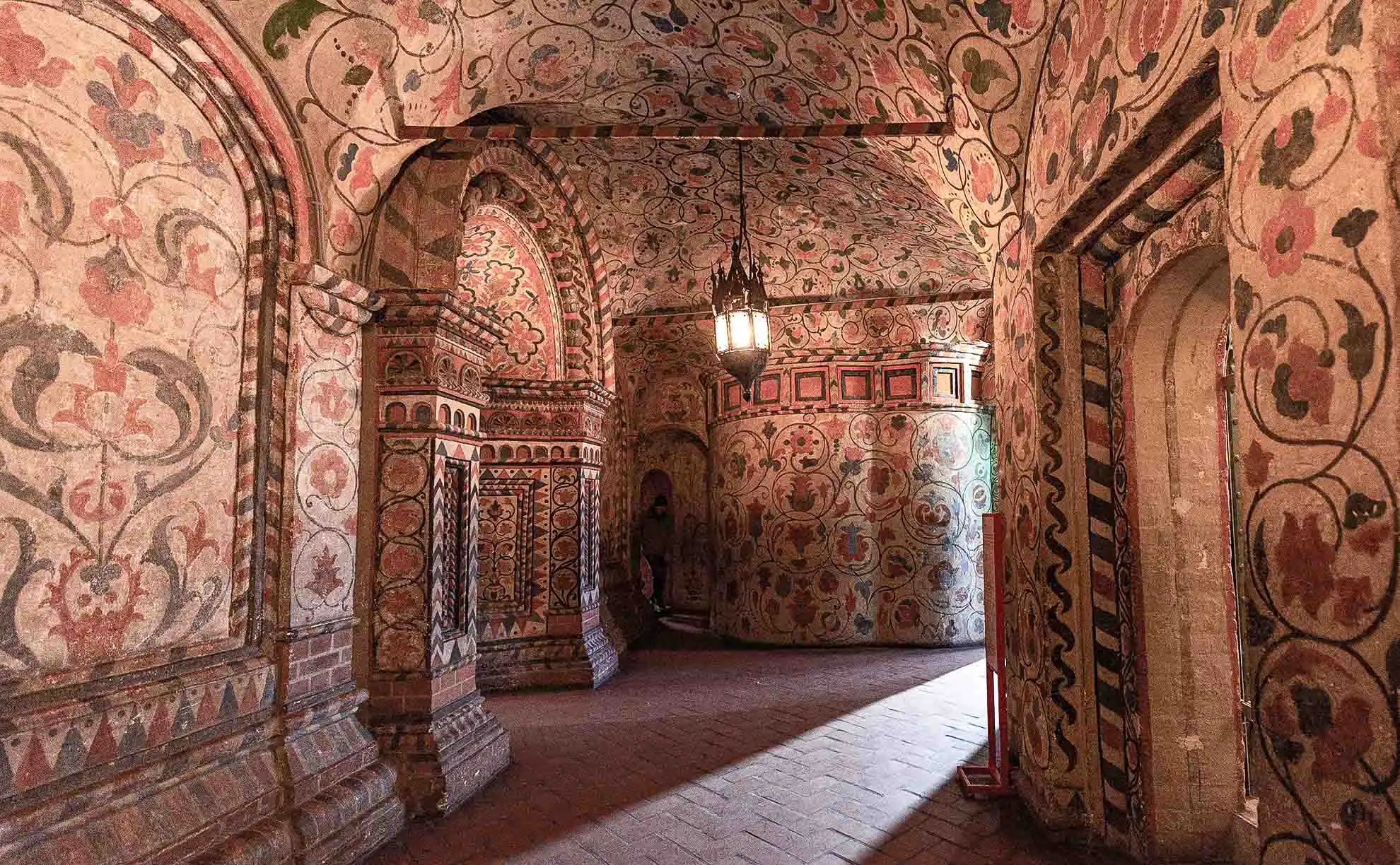 Interior de uma galeria interna estreita da Catedral da Intercessão da Santíssima Mãe de Deus sobre o Fosso, conhecida como Catedral de São Basílio, em Moscou. Os tetos em abóbada arredondada e as maciças paredes de tijolos são completamente cobertos por pinturas do século XVII com ornamentos florais, volutas e flores em tons de vermelho-alaranjado, verde e azul. No piso de tijolos há azulejos retangulares e, ao fundo do corredor, uma luminária escura de ferro forjado pende do teto, criando uma iluminação quente e noturna no interior antigo.