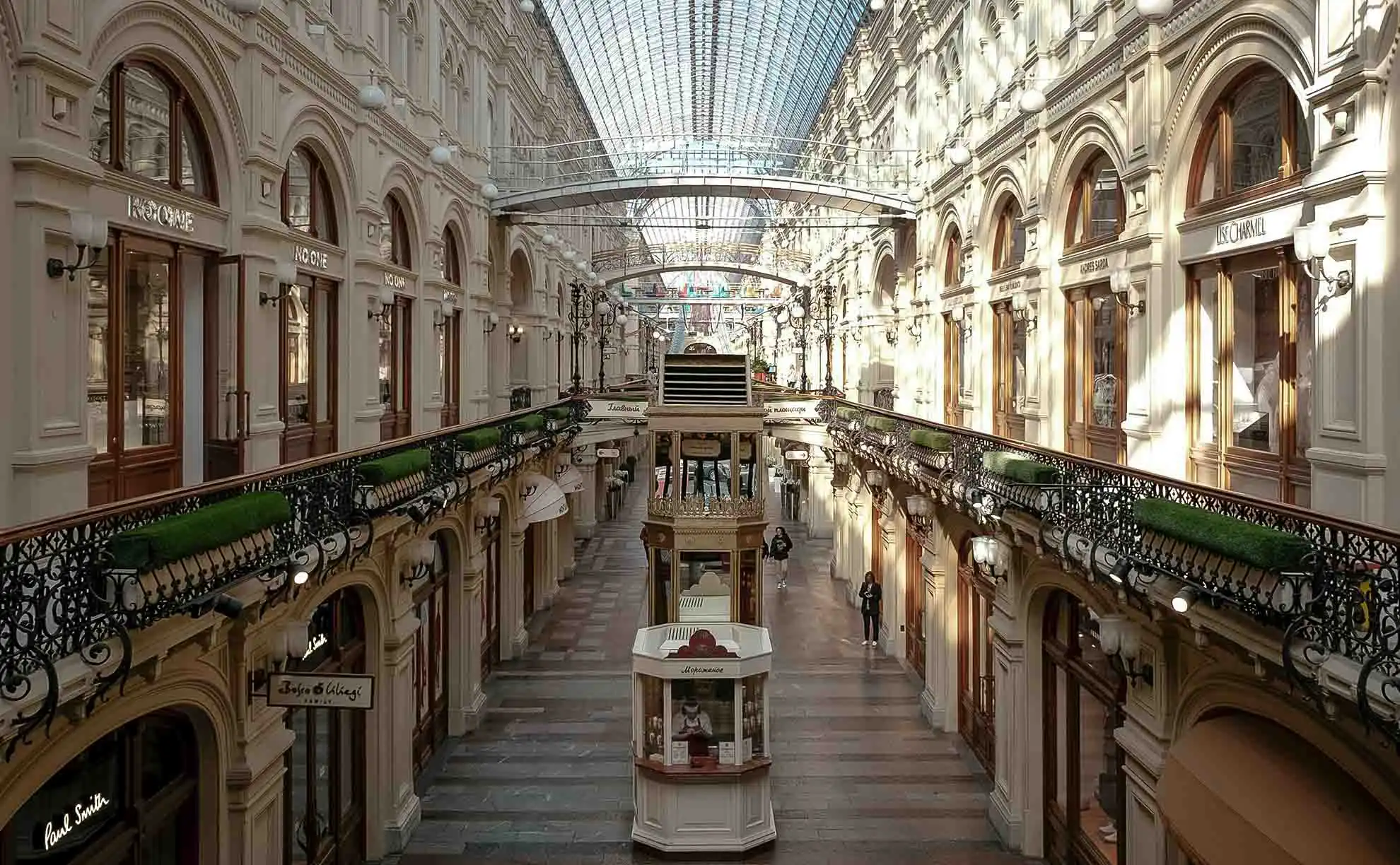 Fachada da Loja de Departamentos GUM em Moscou vista a partir da Praça Vermelha em um dia ensolarado de verão. O monumental edifício de três andares feito de pedra clara é decorado com numerosos arcos entalhados, colunas e padrões, e seu telhado é coroado por duas torres altas e simétricas com pináculos. Diante do prédio, alguns pedestres caminham pelo paralelepípedo, com duas jovens de vestidos de verão olhando para os celulares em primeiro plano, sob um céu azul limpo.