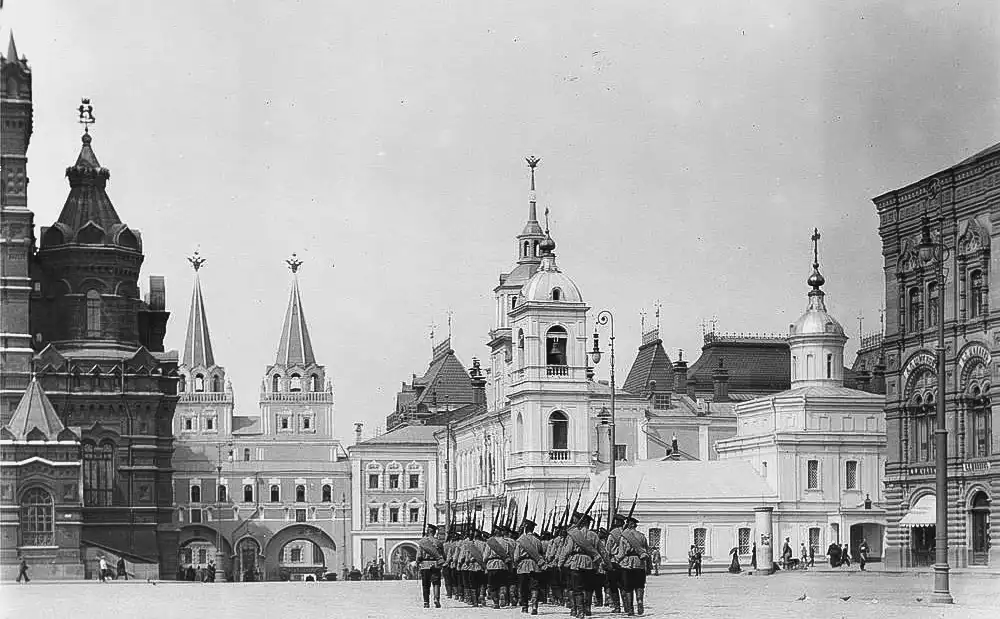 Fotografia histórica em preto e branco do início do século XX, mostrando a vista da Catedral do Ícone de Kazan da Mãe de Deus na Praça Vermelha em Moscou. Pelo calçamento da praça marcha uma fileira de soldados de uniforme militar e quepes, carregando rifles nos ombros. Ao fundo fica o edifício branco de dois andares da catedral com uma torre sineira e uma cúpula. À esquerda, avistam-se as Portas da Ressurreição com duas torres pontiagudas e a fachada escura do Museu Histórico Estatal, à direita estende-se o edifício das Galerias Comerciais Superiores.