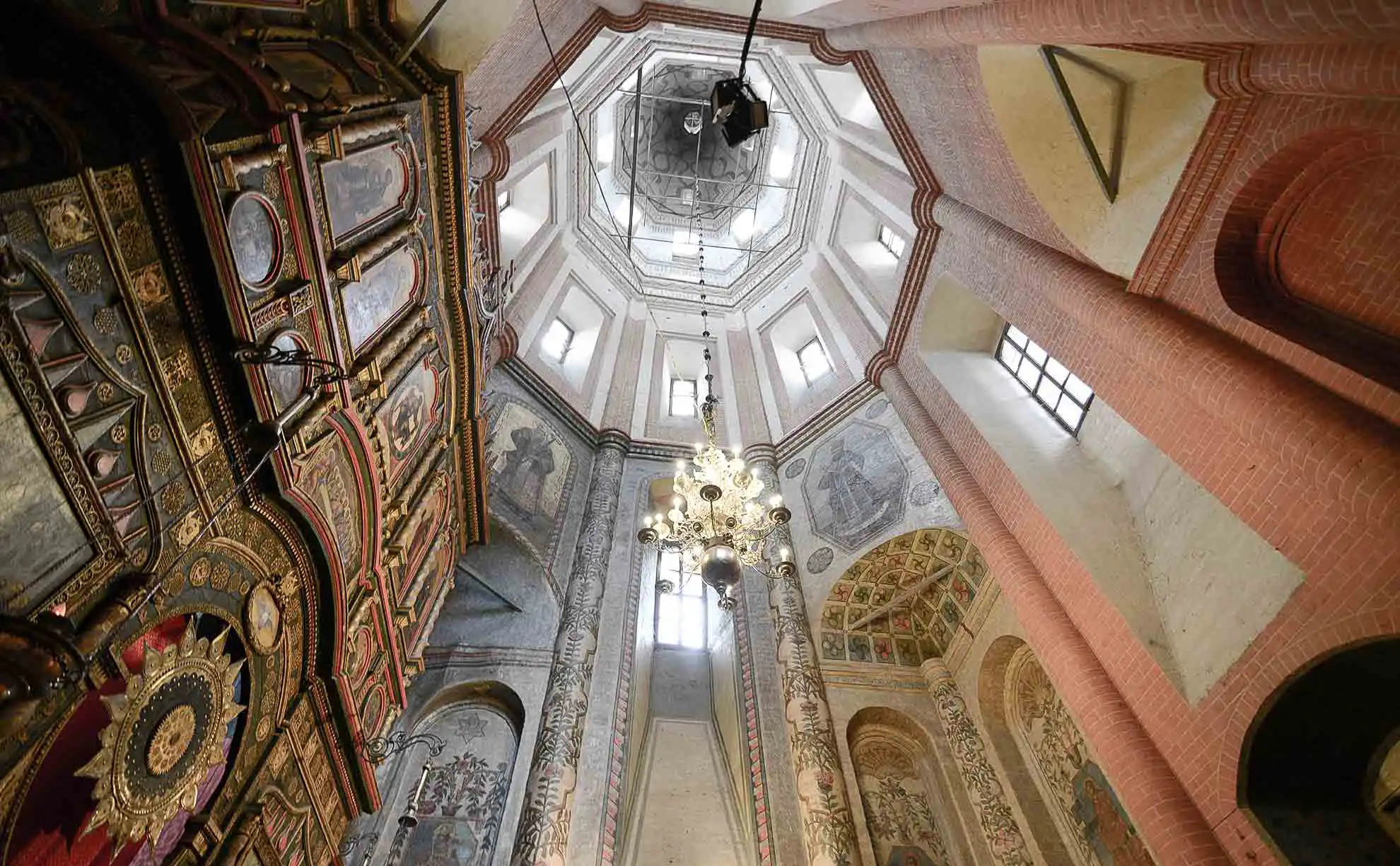 Vista geral da Catedral da Intercessão da Santíssima Mãe de Deus sobre o Fosso, conhecida como Catedral de São Basílio, a partir da Descida de Vassilievsky em Moscou. O monumento arquitetônico de tijolos vermelhos tem uma composição cêntrica com uma alta torre de tenda no centro e cúpulas bulbosas coloridas ao seu redor com padrões retorcidos em verde, azul, amarelo e vermelho. Diante do templo, no paralelepípedo da Praça Vermelha, caminhham dezenas de turistas com roupas de verão, à esquerda avisa-se o edifício amarelo das Antigas Fileiras Comerciais Médias e acima estende-se um céu azul vibrante com nuvens brancas leves.