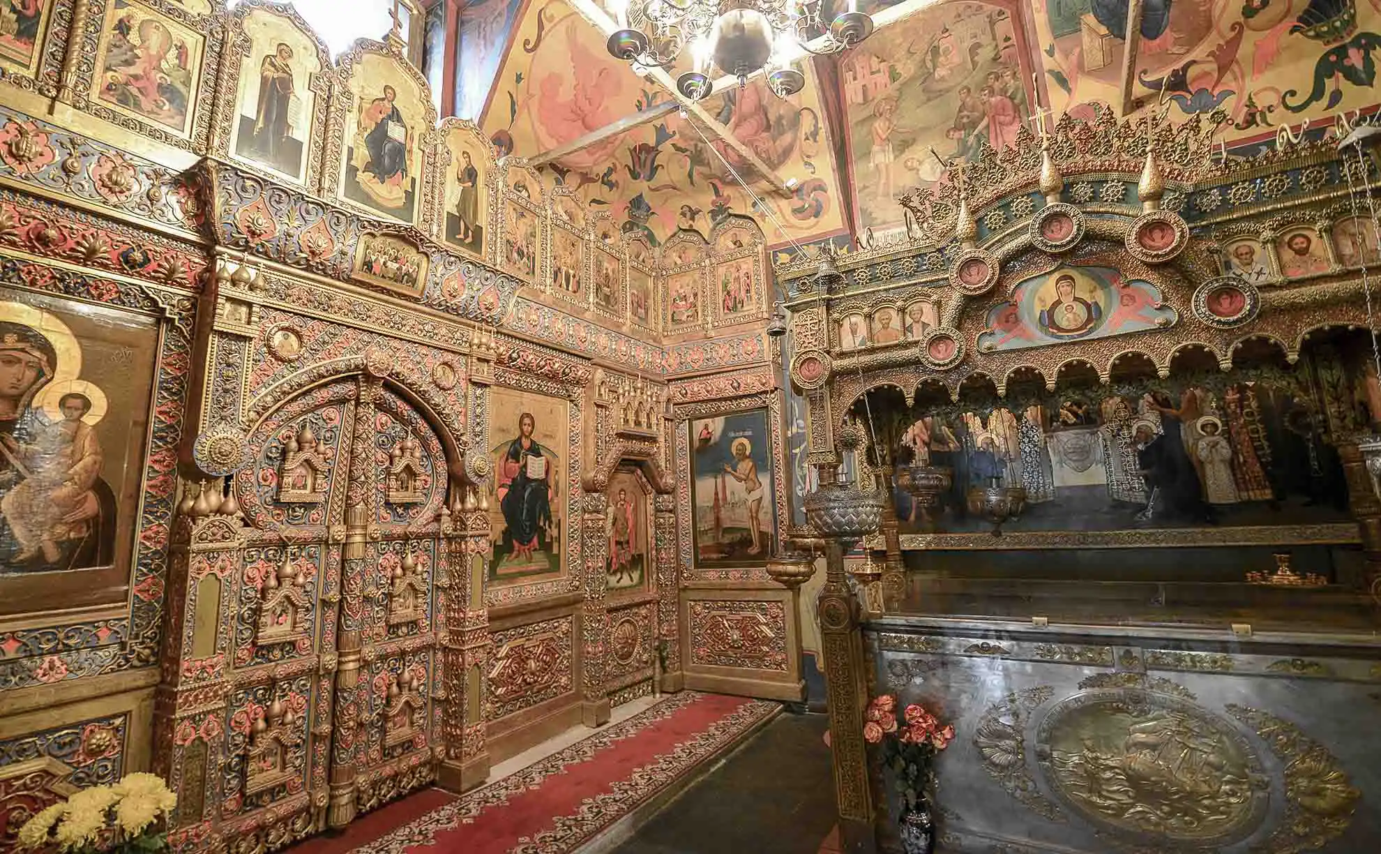 Vista geral da Catedral da Intercessão da Santíssima Mãe de Deus sobre o Fosso, conhecida como Catedral de São Basílio, a partir da Descida de Vassilievsky em Moscou. O monumento arquitetônico de tijolos vermelhos tem uma composição cêntrica com uma alta torre de tenda no centro e cúpulas bulbosas coloridas ao seu redor com padrões retorcidos em verde, azul, amarelo e vermelho. Diante do templo, no paralelepípedo da Praça Vermelha, caminhham dezenas de turistas com roupas de verão, à esquerda avisa-se o edifício amarelo das Antigas Fileiras Comerciais Médias e acima estende-se um céu azul vibrante com nuvens brancas leves.