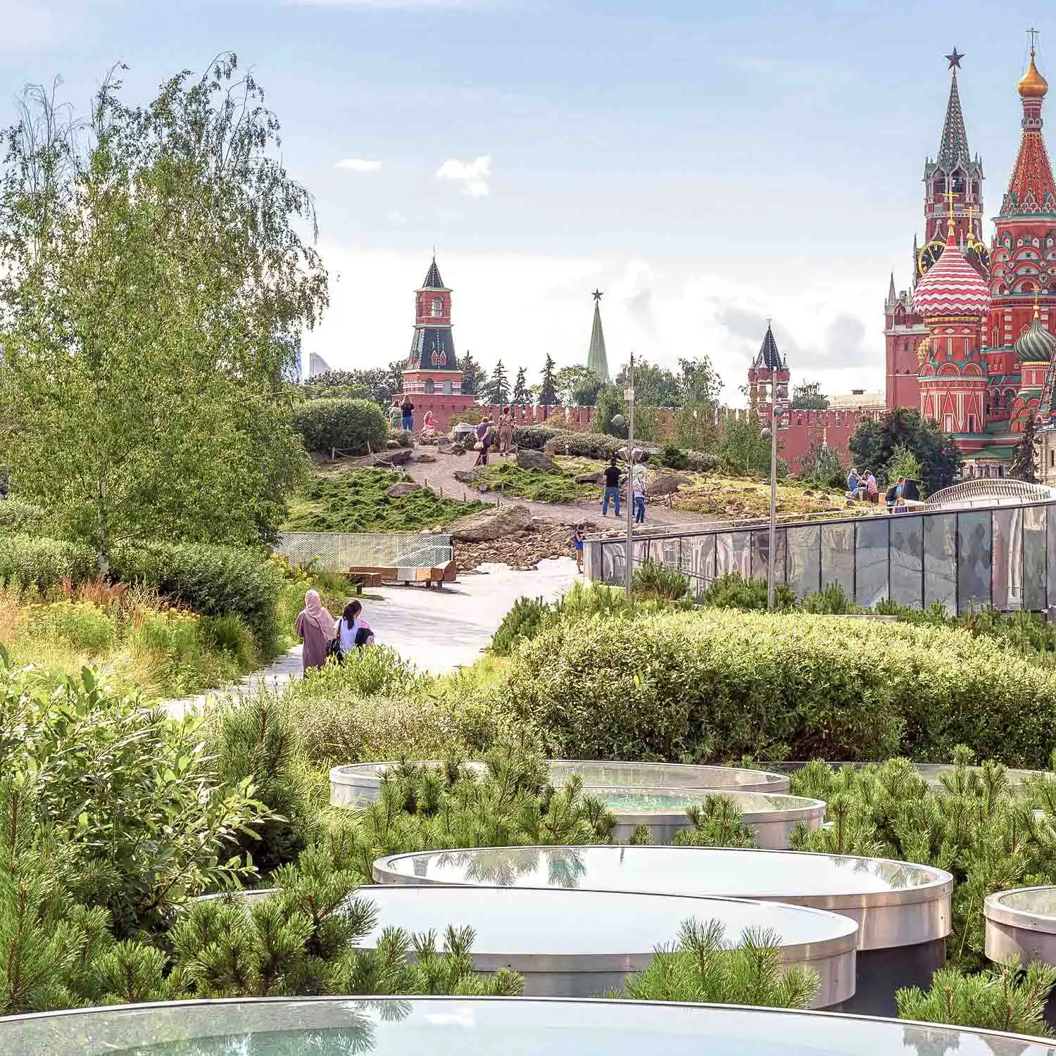 Área paisagística vegetal do Parque Zaryadye em Moscou. Em primeiro plano estão as janelas redondas de vidro dos pavilhões subterrâneos, cercadas por arbustos verdes de agulhas e grama. Uma trilha de pedestres segue para o interior da colina, onde pessoas caminham. Ao fundo, atrás de uma cerca baixa de vidro, abre-se o panorama para a muralha de tijolos vermelhos do Kremlin de Moscou, suas torres e as cúpulas coloridas da Catedral da Intercessão da Santíssima Mãe de Deus sobre o Fosso, conhecida como Catedral de São Basílio.