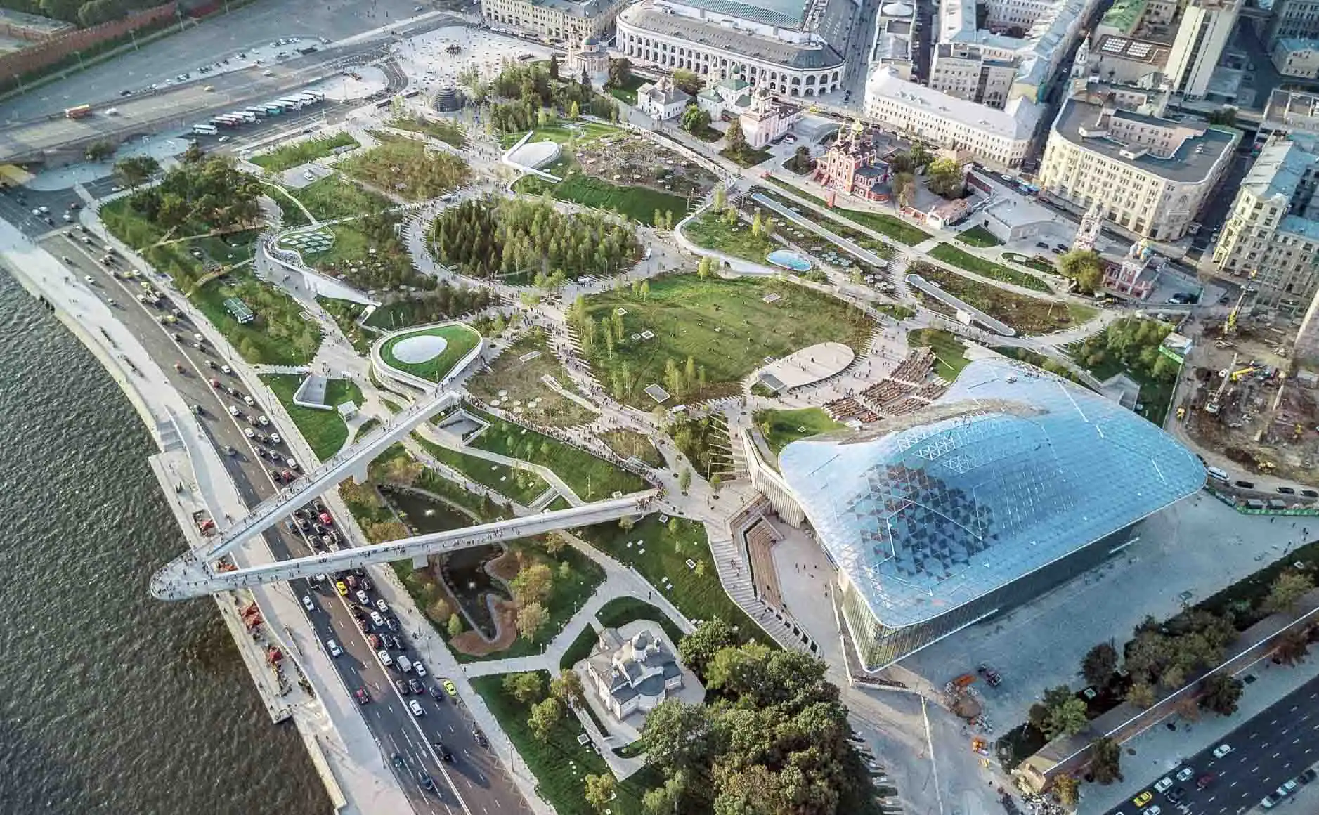 Área paisagística vegetal do Parque Zaryadye em Moscou. Em primeiro plano estão as janelas redondas de vidro dos pavilhões subterrâneos, cercadas por arbustos verdes de agulhas e grama. Uma trilha de pedestres segue para o interior da colina, onde pessoas caminham. Ao fundo, atrás de uma cerca baixa de vidro, abre-se o panorama para a muralha de tijolos vermelhos do Kremlin de Moscou, suas torres e as cúpulas coloridas da Catedral da Intercessão da Santíssima Mãe de Deus sobre o Fosso, conhecida como Catedral de São Basílio.