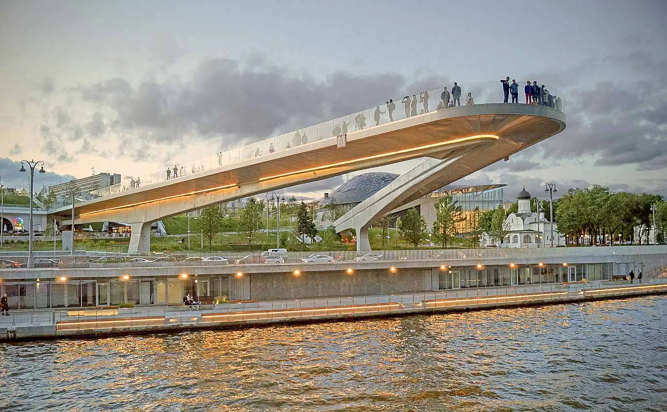Área paisagística vegetal do Parque Zaryadye em Moscou. Em primeiro plano estão as janelas redondas de vidro dos pavilhões subterrâneos, cercadas por arbustos verdes de agulhas e grama. Uma trilha de pedestres segue para o interior da colina, onde pessoas caminham. Ao fundo, atrás de uma cerca baixa de vidro, abre-se o panorama para a muralha de tijolos vermelhos do Kremlin de Moscou, suas torres e as cúpulas coloridas da Catedral da Intercessão da Santíssima Mãe de Deus sobre o Fosso, conhecida como Catedral de São Basílio.