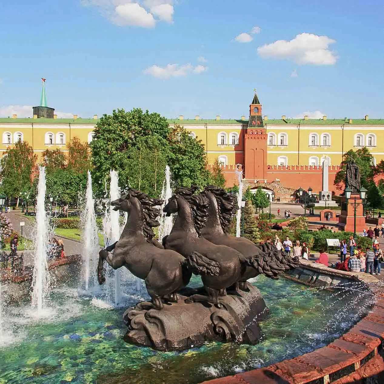 Vista pitoresca da fonte no Jardim Alexandre do Kremlin de Moscou em um dia ensolarado. No centro de uma piscina circular com jatos de água, ergue-se um maciço grupo escultórico em bronze escuro de quatro cavalos empinados em poses dinâmicas. Ao fundo, atrás das copas verdes das árvores, estende-se a fachada amarela do Edifício do Arsenal do Kremlin e a Torre Média do Arsenal sob um céu azul com nuvens brancas.