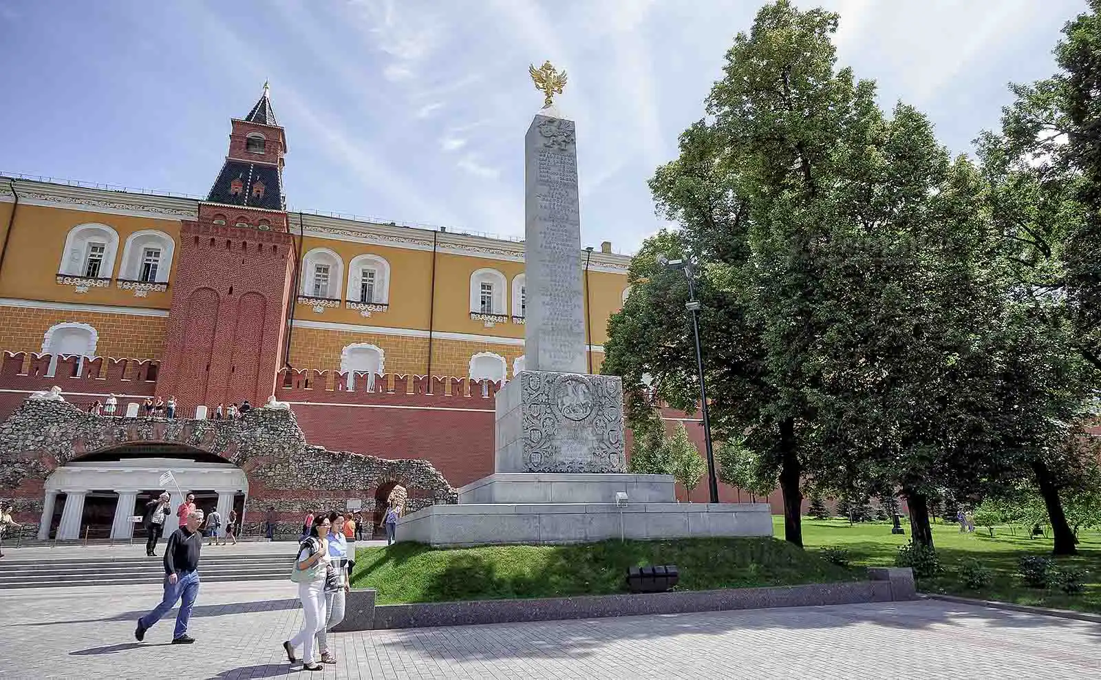 Vista pitoresca da fonte no Jardim Alexandre do Kremlin de Moscou em um dia ensolarado. No centro de uma piscina circular com jatos de água, ergue-se um maciço grupo escultórico em bronze escuro de quatro cavalos empinados em poses dinâmicas. Ao fundo, atrás das copas verdes das árvores, estende-se a fachada amarela do Edifício do Arsenal do Kremlin e a Torre Média do Arsenal sob um céu azul com nuvens brancas.