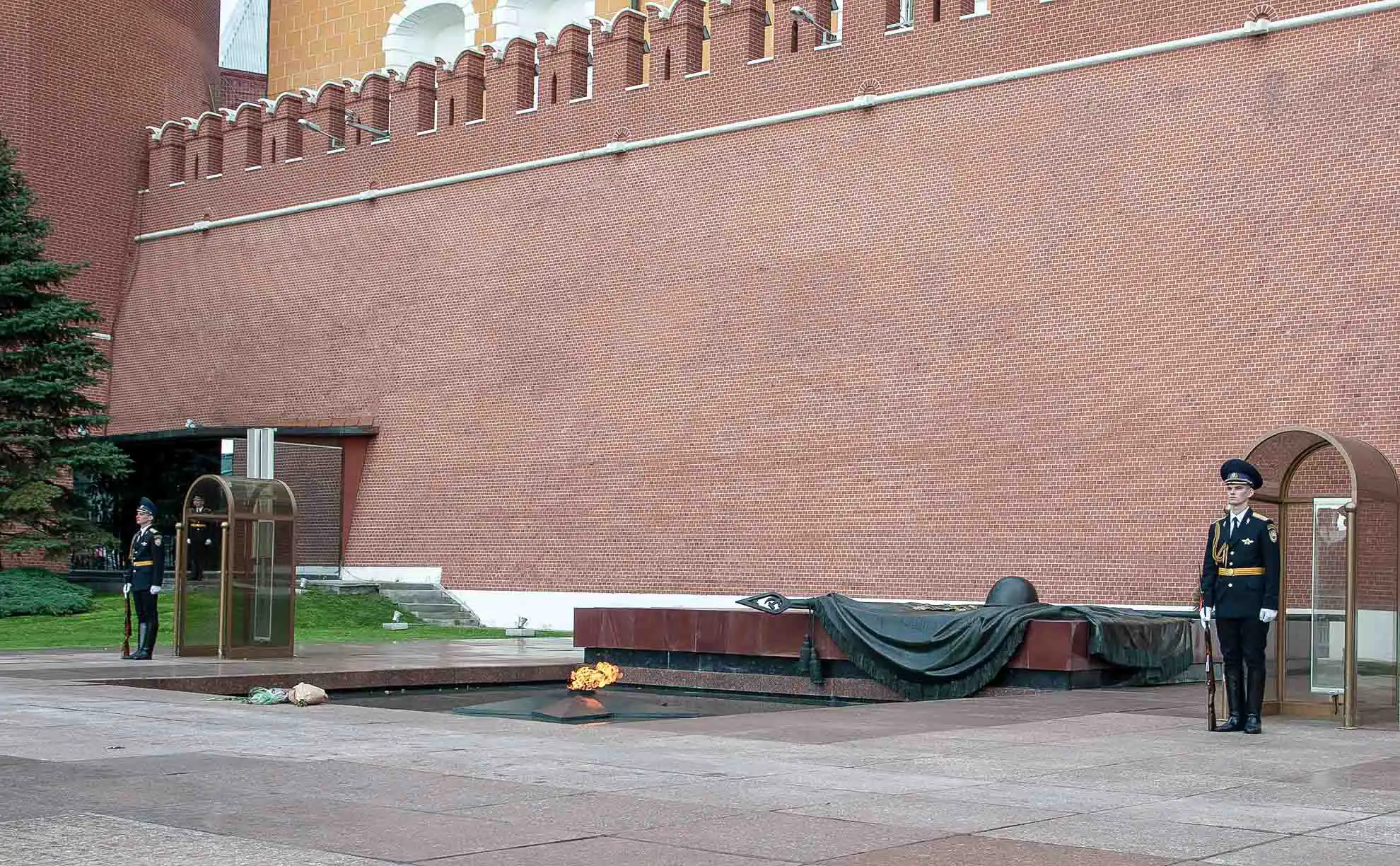 Vista pitoresca da fonte no Jardim Alexandre do Kremlin de Moscou em um dia ensolarado. No centro de uma piscina circular com jatos de água, ergue-se um maciço grupo escultórico em bronze escuro de quatro cavalos empinados em poses dinâmicas. Ao fundo, atrás das copas verdes das árvores, estende-se a fachada amarela do Edifício do Arsenal do Kremlin e a Torre Média do Arsenal sob um céu azul com nuvens brancas.