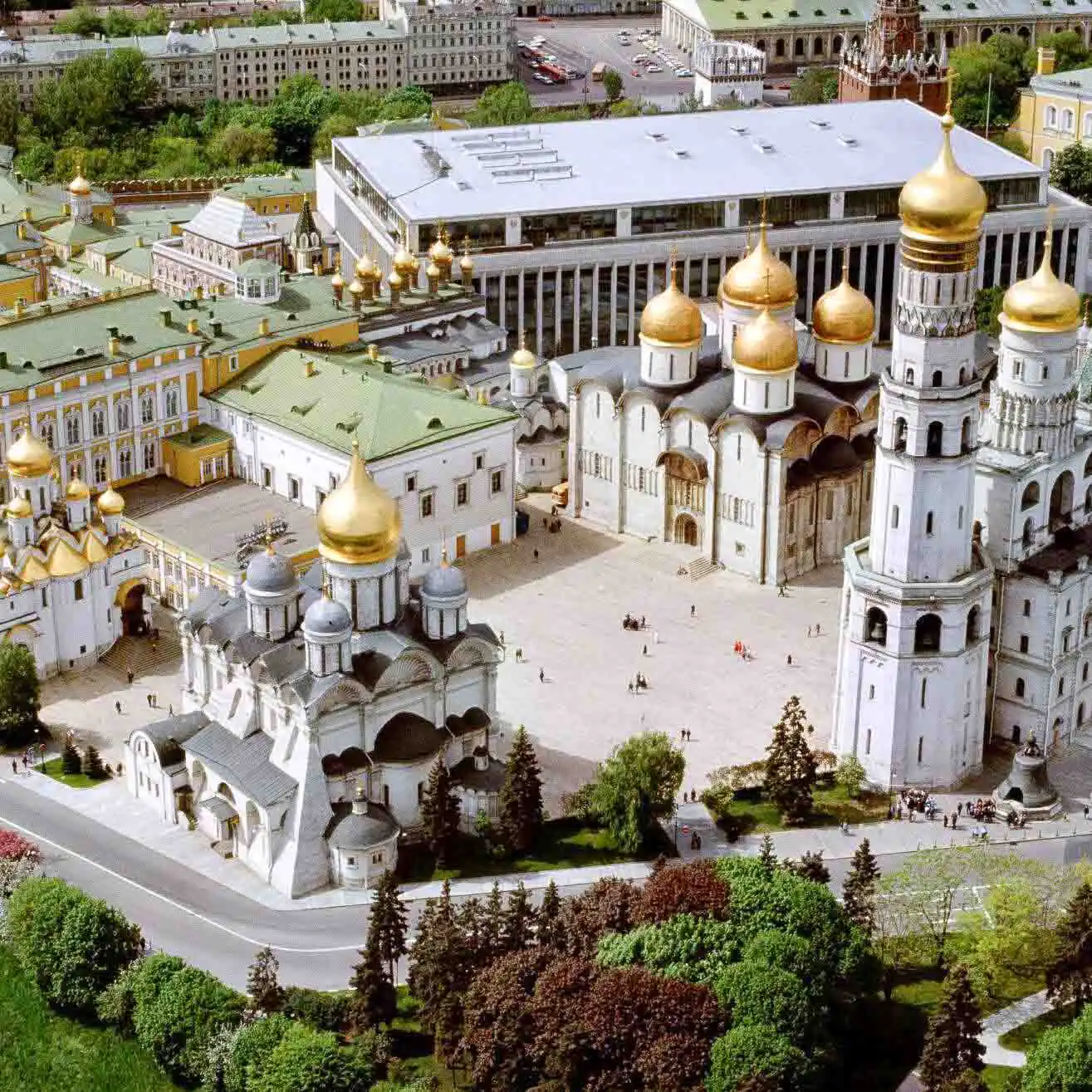 Vista aérea da Praça das Catedrais do Kremlin de Moscou em um dia ensolarado. No centro ergue-se a torre branca multinível do Sino de Ivan, o Grande, com uma grande cúpula dourada. Ao seu redor localizam-se os maciços edifícios das catedrais com múltiplas cúpulas douradas e prateadas, incluindo as catedrais da Dormição e do Arcanjo. Em primeiro plano vê-se o calçamento de pedra cinza e ao fundo, atrás das árvores do parque, corre o rio Moscou sob o céu azul.