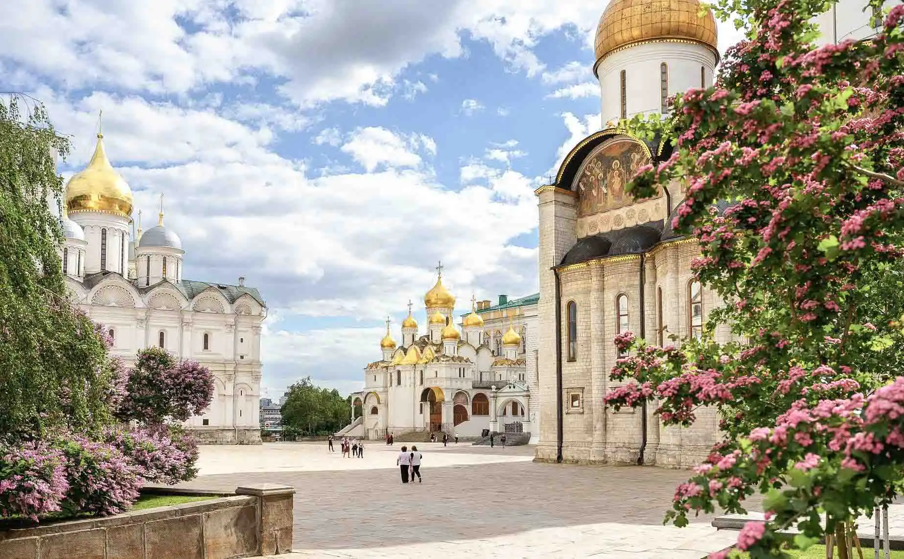 Vista aérea da Praça das Catedrais do Kremlin de Moscou em um dia ensolarado. No centro ergue-se a torre branca multinível do Sino de Ivan, o Grande, com uma grande cúpula dourada. Ao seu redor localizam-se os maciços edifícios das catedrais com múltiplas cúpulas douradas e prateadas, incluindo as catedrais da Dormição e do Arcanjo. Em primeiro plano vê-se o calçamento de pedra cinza e ao fundo, atrás das árvores do parque, corre o rio Moscou sob o céu azul.