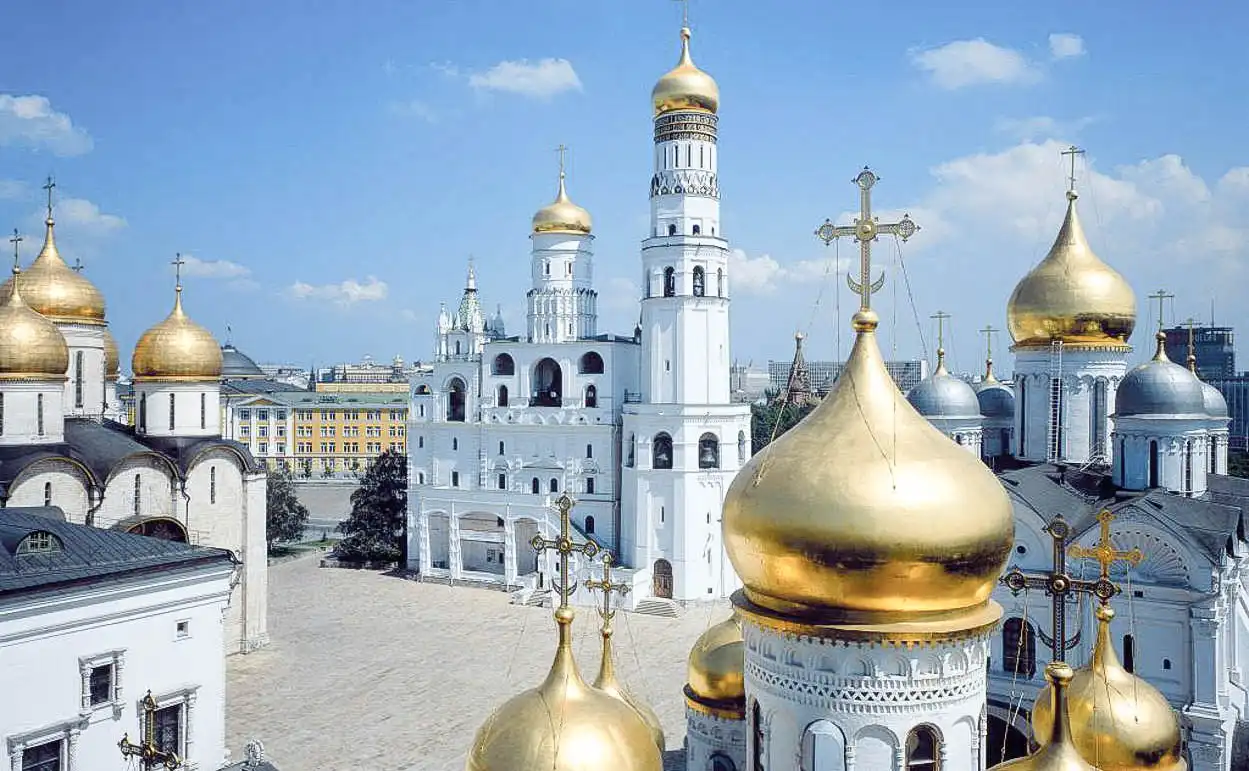 Vista aérea da Praça das Catedrais do Kremlin de Moscou em um dia ensolarado. No centro ergue-se a torre branca multinível do Sino de Ivan, o Grande, com uma grande cúpula dourada. Ao seu redor localizam-se os maciços edifícios das catedrais com múltiplas cúpulas douradas e prateadas, incluindo as catedrais da Dormição e do Arcanjo. Em primeiro plano vê-se o calçamento de pedra cinza e ao fundo, atrás das árvores do parque, corre o rio Moscou sob o céu azul.