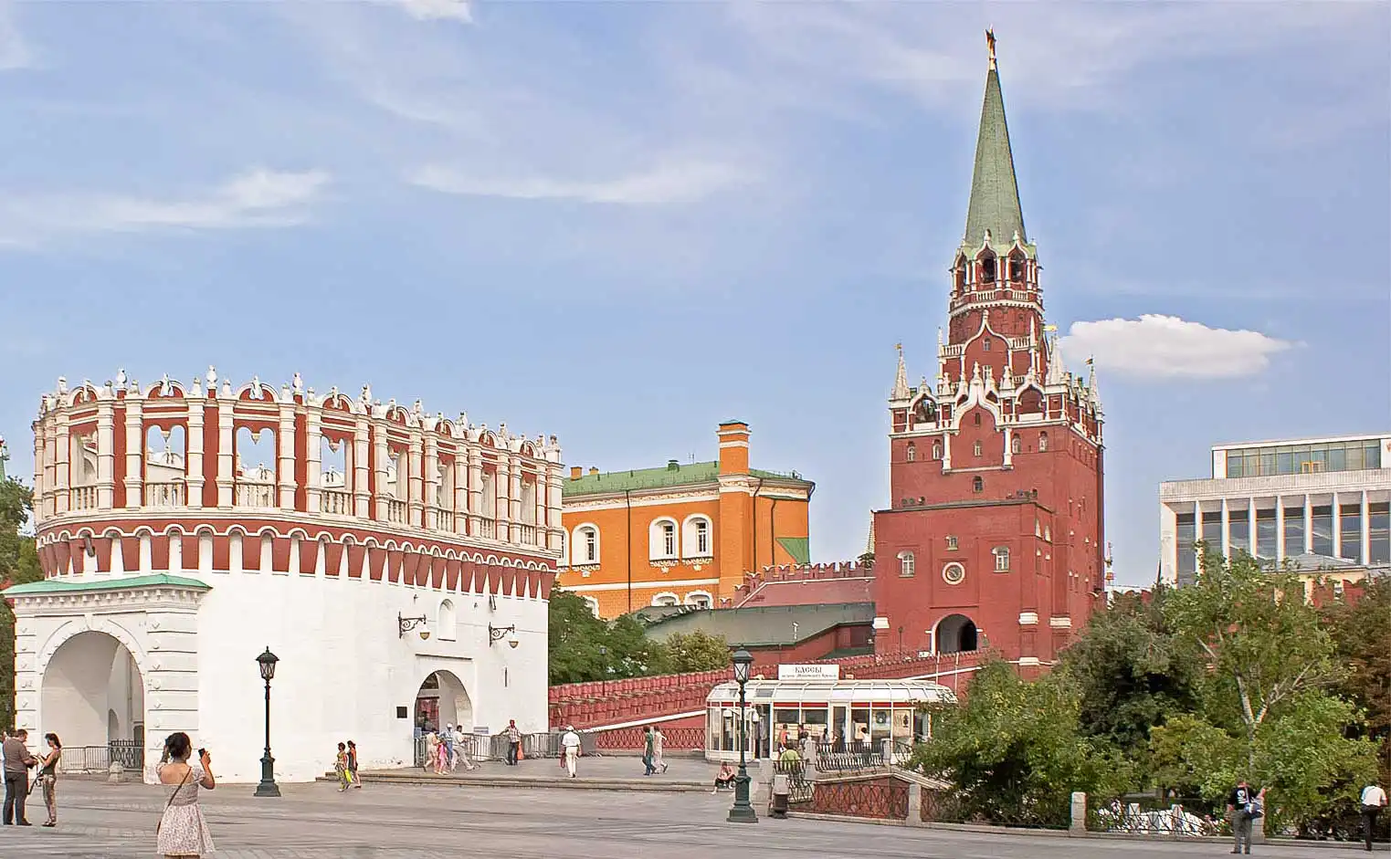 Praça em frente à Torre Kutafiya do Kremlin de Moscou sob o céu nublado. À esquerda está a Torre Kutafiya, branca e arredondada, com topo denteado decorativo em vermelho e branco e passagens em arco. À direita, atrás da ponte, ergue-se a alta Torre Troitskaya de tijolos vermelhos com uma agulha e telhado em estilo tenda verde. Em primeiro plano, pessoas caminham pela praça pavimentada, com um homem à esquerda fotografando uma pessoa sentada no chão.