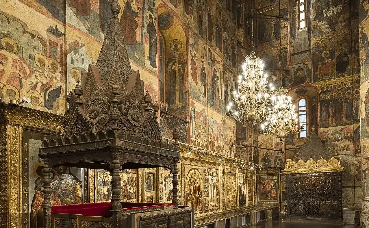 Fachada de pedra branca da Catedral da Dormição do Kremlin de Moscou em um dia ensolarado e claro. O maciço edifício retangular é decorado com arcos semicirculares, janelas altas e estreitas e coroado por cinco grandes cúpulas douradas em formato de capacete com cruzes de ferro forjado. Acima da entrada principal, avista-se um grande afresco retratando a Mãe de Deus, diante da catedral estende-se a Praça das Catedrais vazia e, à direita, veem-se as paredes brancas entalhadas do Palácio de Facetas sob o céu azul com nuvens leves.