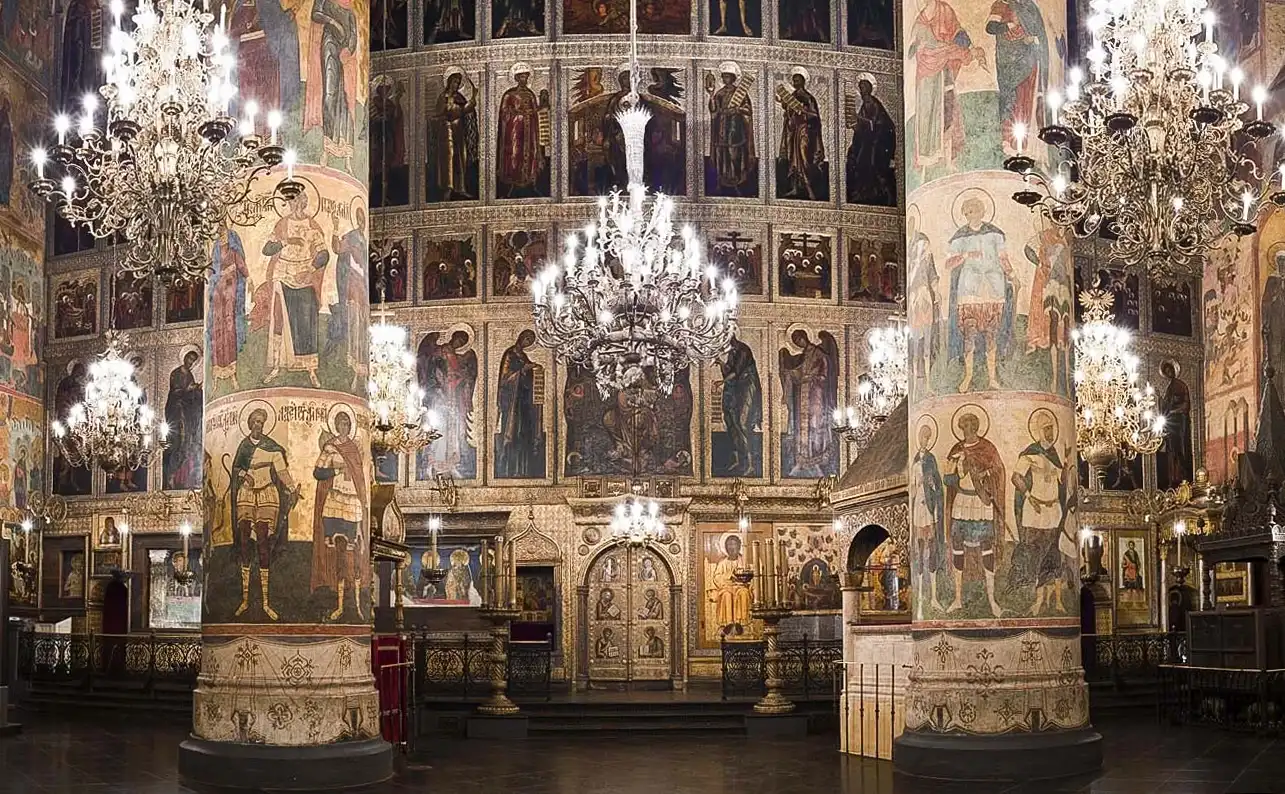 Fachada de pedra branca da Catedral da Dormição do Kremlin de Moscou em um dia ensolarado e claro. O maciço edifício retangular é decorado com arcos semicirculares, janelas altas e estreitas e coroado por cinco grandes cúpulas douradas em formato de capacete com cruzes de ferro forjado. Acima da entrada principal, avista-se um grande afresco retratando a Mãe de Deus, diante da catedral estende-se a Praça das Catedrais vazia e, à direita, veem-se as paredes brancas entalhadas do Palácio de Facetas sob o céu azul com nuvens leves.