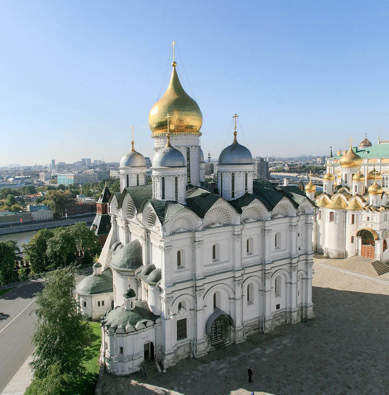 Fachada de pedra branca da Catedral de São Miguel Arcanjo do Kremlin de Moscou em um dia ensolarado e claro. O edifício retangular de dois andares é decorado com pilastras, arcos semicirculares com ornamentos em relevo entalhado em formato de conchas e coroado por uma grande cúpula dourada e quatro cúpulas prateadas menores. Ao fundo, à direita, avista-se a fachada clara da Catedral da Anunciação com suas múltiplas cúpulas douradas, e diante da catedral estende-se a praça vazia de piso cinza sob o céu azul limpo.