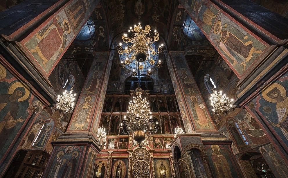 Fachada de pedra branca da Catedral de São Miguel Arcanjo do Kremlin de Moscou em um dia ensolarado e claro. O edifício retangular de dois andares é decorado com pilastras, arcos semicirculares com ornamentos em relevo entalhado em formato de conchas e coroado por uma grande cúpula dourada e quatro cúpulas prateadas menores. Ao fundo, à direita, avista-se a fachada clara da Catedral da Anunciação com suas múltiplas cúpulas douradas, e diante da catedral estende-se a praça vazia de piso cinza sob o céu azul limpo.