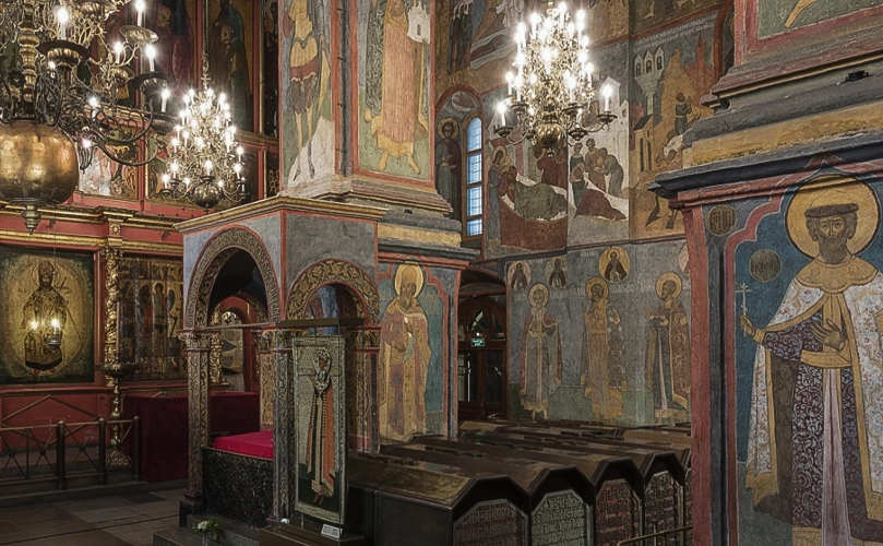 Vista da necrópole histórica no interior da Catedral de São Miguel Arcanjo do Kremlin de Moscou. No centro e ao longo das paredes do recinto estão instaladas longas fileiras de lápides retangulares idênticas de governantes, feitas de pedra escura com inscrições entalhadas nas extremidades. Nas paredes e na maciça coluna de suporte veem-se afrescos antigos com imagens de santos, à esquerda junto ao iconóstase dourado fica o Assento Real, e lustres de cristal com velas pendem do teto.