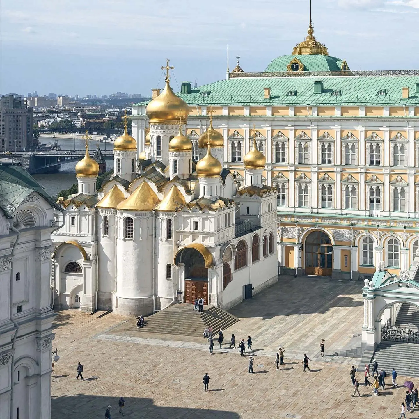 Praça no Kremlin de Moscou com vista para a Catedral da Anunciação de pedra branca em um dia ensolarado. O edifício da catedral possui uma arquitetura escalonada complexa com galerias em arco abertas e é coroado por uma grande cúpula dourada e quatro cúpulas prateadas menores. Em primeiro plano, à direita, fica o edifício branco do Palácio de Facetas com sua fachada de pedra lapidada e, ao fundo, ergue-se o maciço corpo amarelo do Grande Palácio do Kremlin sob o céu azul com nuvens leves.
