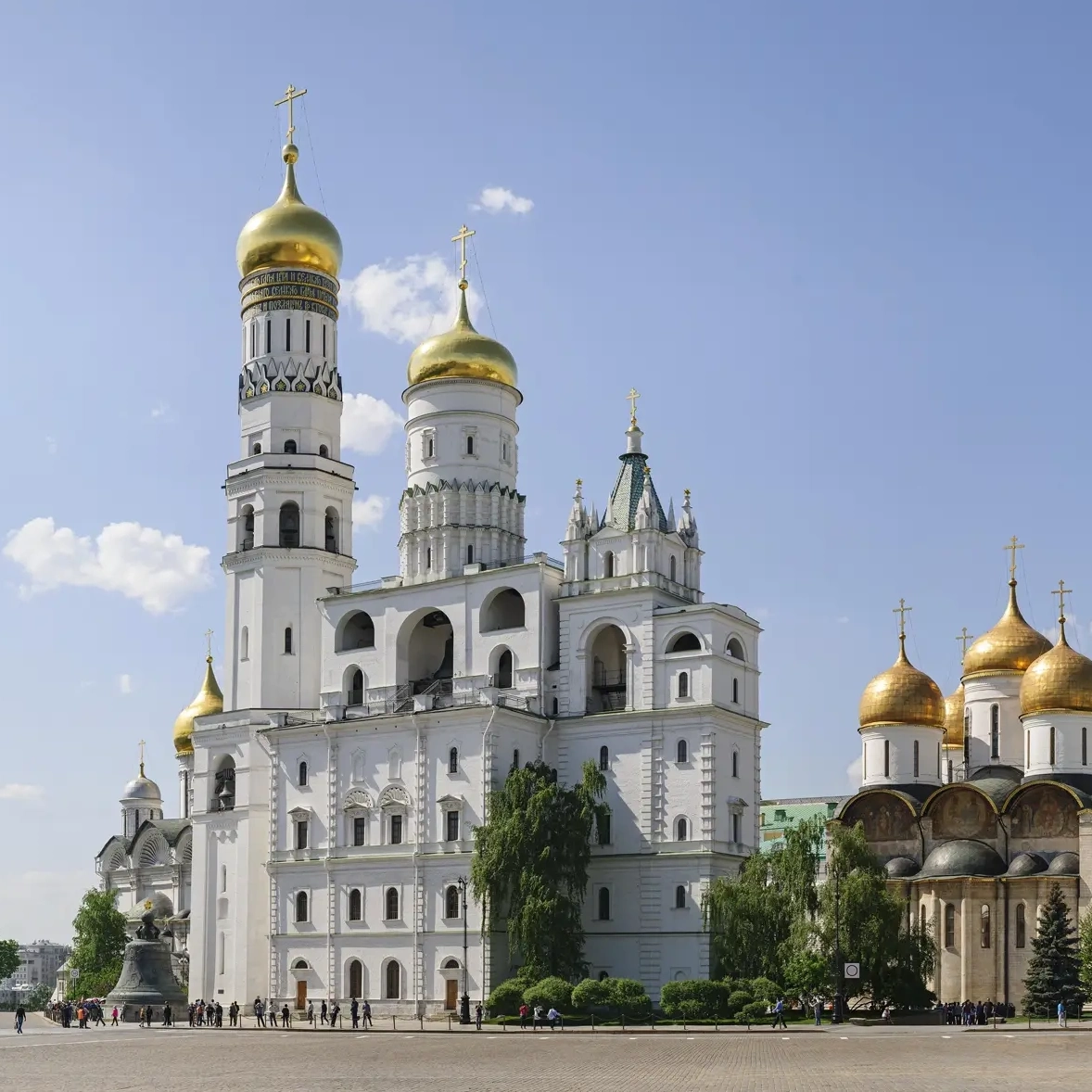 Conjunto arquitetônico da Torre do Sino de Ivan, o Grande, na Praça das Catedrais do Kremlin de Moscou em um dia ensolarado de verão. À esquerda ergue-se o pilar octogonal branco da torre com cúpula dourada, no centro localiza-se a Torre Sineira da Assunção de quatro níveis com aberturas em arco e sinos, e à direita anexa-se o Anexo de Filareto com topo em tenda e cúpula de ouro. Em primeiro plano, numerosos turistas caminham pela esplanada pavimentada sob um céu azul com nuvens cúmulas brancas.