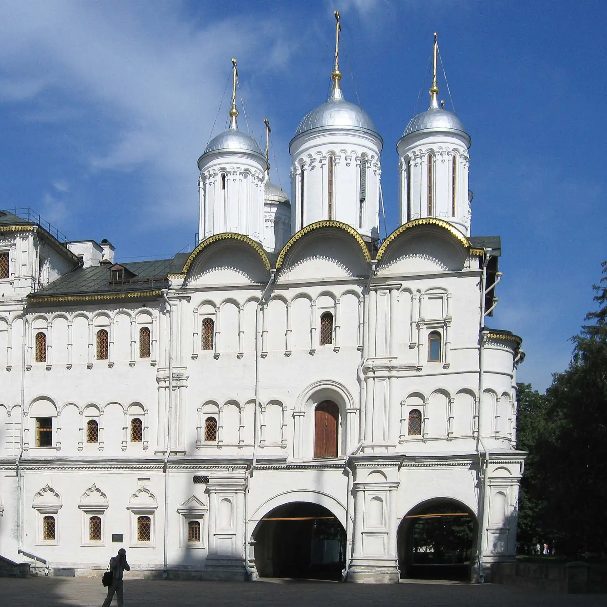 Fachada das Câmaras Patriarcais com a Igreja dos Doze Apóstolos na Praça das Catedrais do Kremlin de Moscou em um dia claro. O monumental edifício branco de três andares com arcos de passagem na base é coroado por cinco cúpulas prateadas sobre altos tambores com cruzes douradas. À esquerda, destaca-se a maciça parede de pedra branca da Catedral da Dormição, enquanto pedestres caminham pela praça pavimentada sob o céu azul.