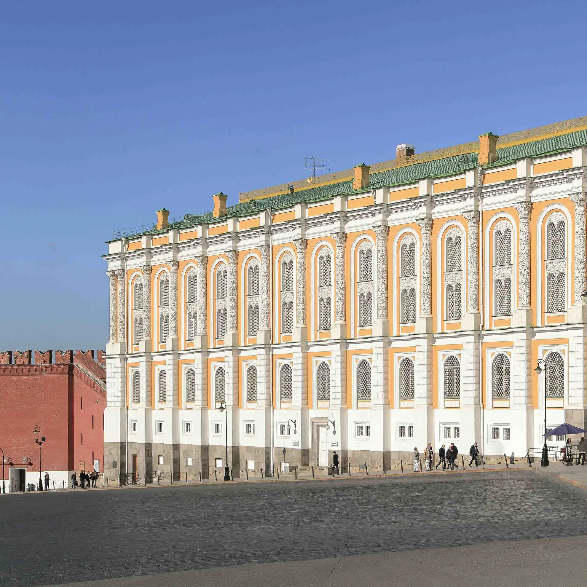 Fachada monumental de dois andares do edifício da Armaria no Kremlin de Moscou em um dia ensolarado. O longo edifício em amarelo e branco no estilo neorrenascentista com telhado verde inclinado exibe fileiras regulares de janelas em arco decoradas com molduras brancas e semicolunas entalhadas. À esquerda, conecta-se a muralha de tijolos vermelhos com ameias e a alta Torre Borovitskaya sob teto cônico verde, enquanto o primeiro plano traz uma avenida asfaltada sob céu azul com nuvens.