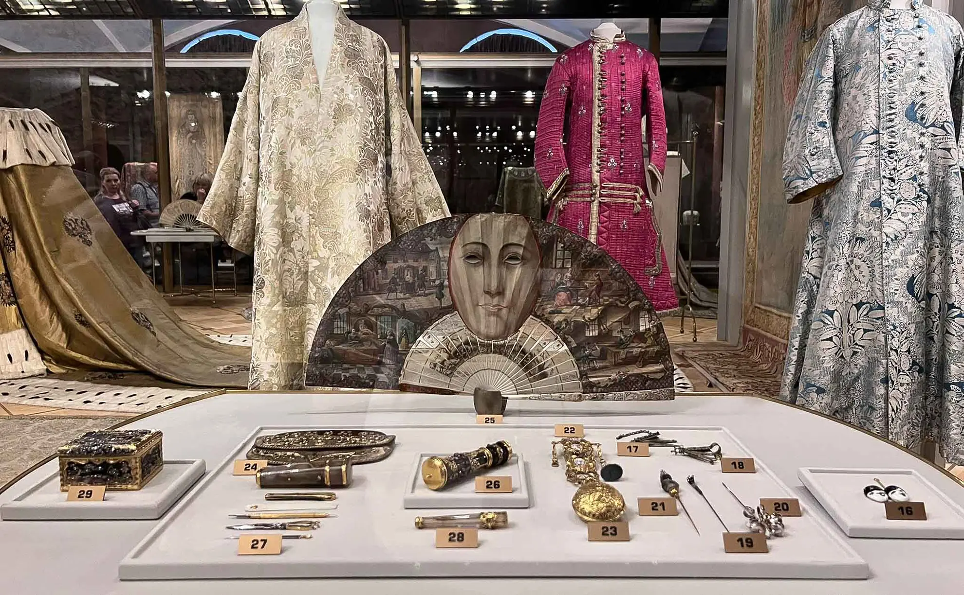 Exposição de vestimentas de corte e acessórios do século XVIII em vitrine de vidro da Armaria do Kremlin. Em primeiro plano, sobre um mostrador branco numerado, descansam relógios de bolso em ouro, caixas de rapé, frascos decorados e um leque semicircular pintado com um rosto feminino. Ao fundo, manequins exibem trajes históricos: um robe de seda clara com bordados prateados, um casaco comprido magenta com botões alinhados e um longo vestido azul estampado.