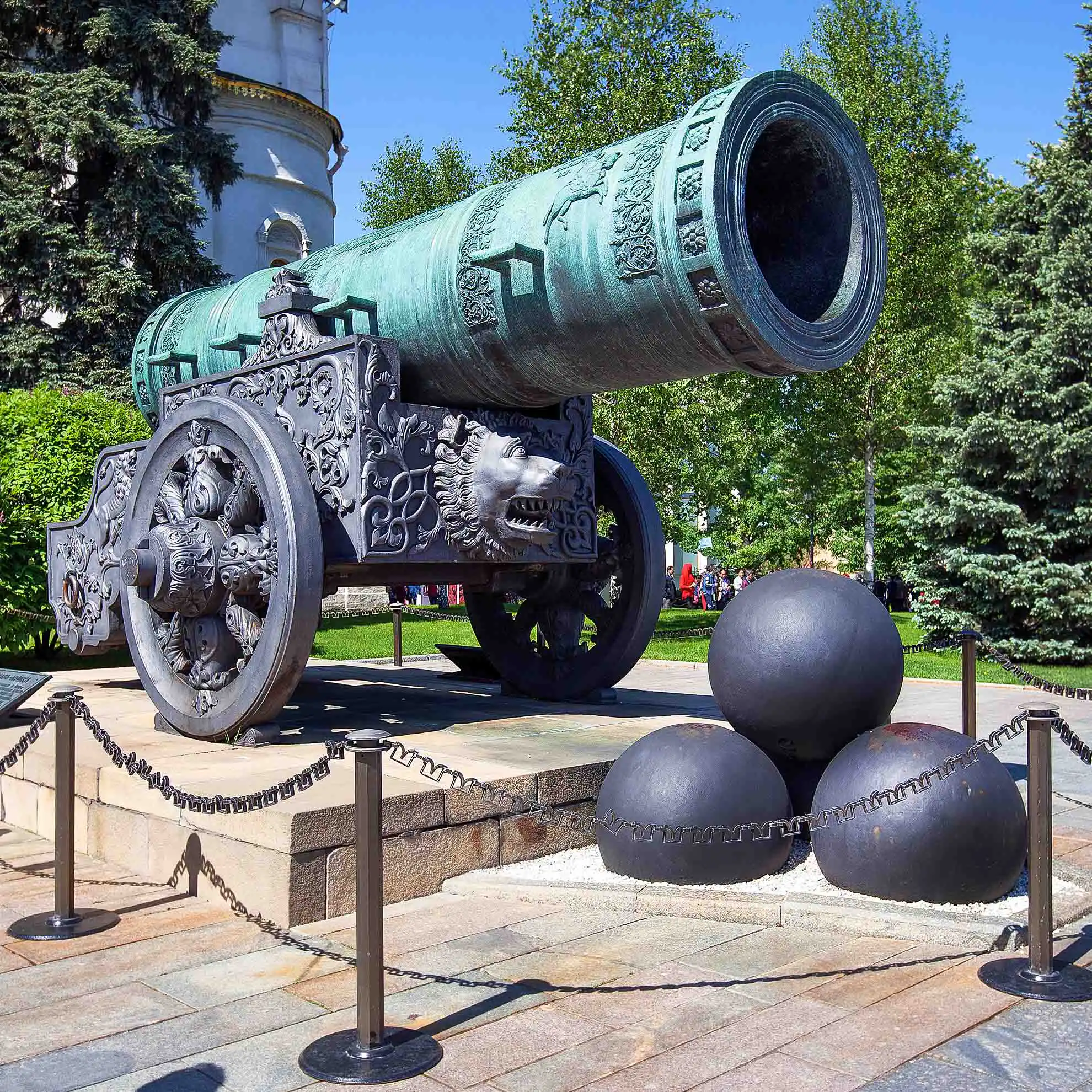 Canhao monumental de bronze historico em tom esverdeado montado sobre uma base de quatro rodas de metal esculpido no Kremlin de Moscou. A enorme boca redonda do cano aponta para o alto em direcao a direita, diante da qual repousam tres imensas esferas de ferro preto em formato de piramide cercadas por uma corrente de protecao. Ao fundo destacam-se arvores verdes e a fachada clara de um edificio historico sob o ceu azul ensolarado.